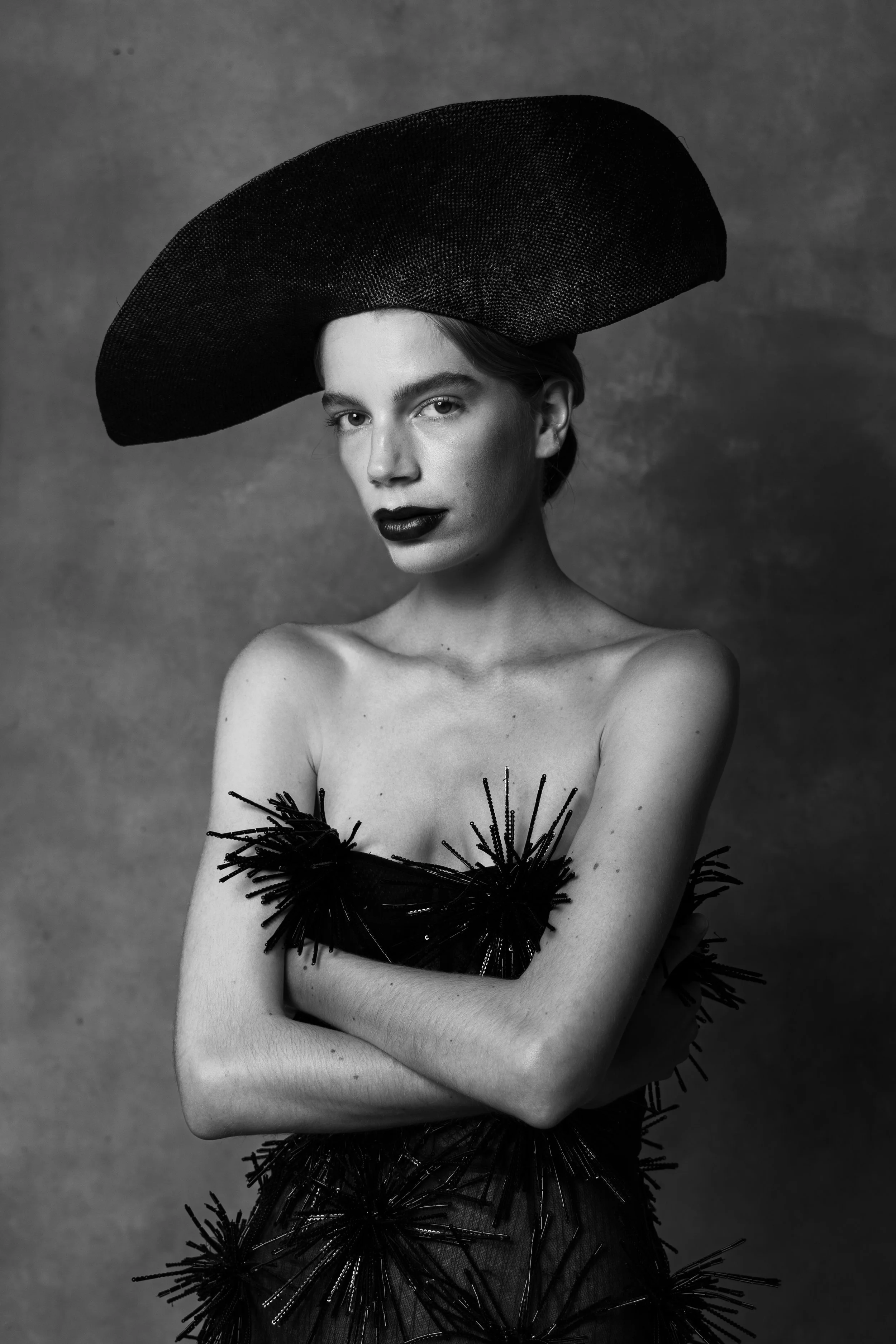 Black and white photo of a woman wearing a wide-brimmed hat and an avant-garde dress with spiky embellishments, arms crossed, against a textured background.