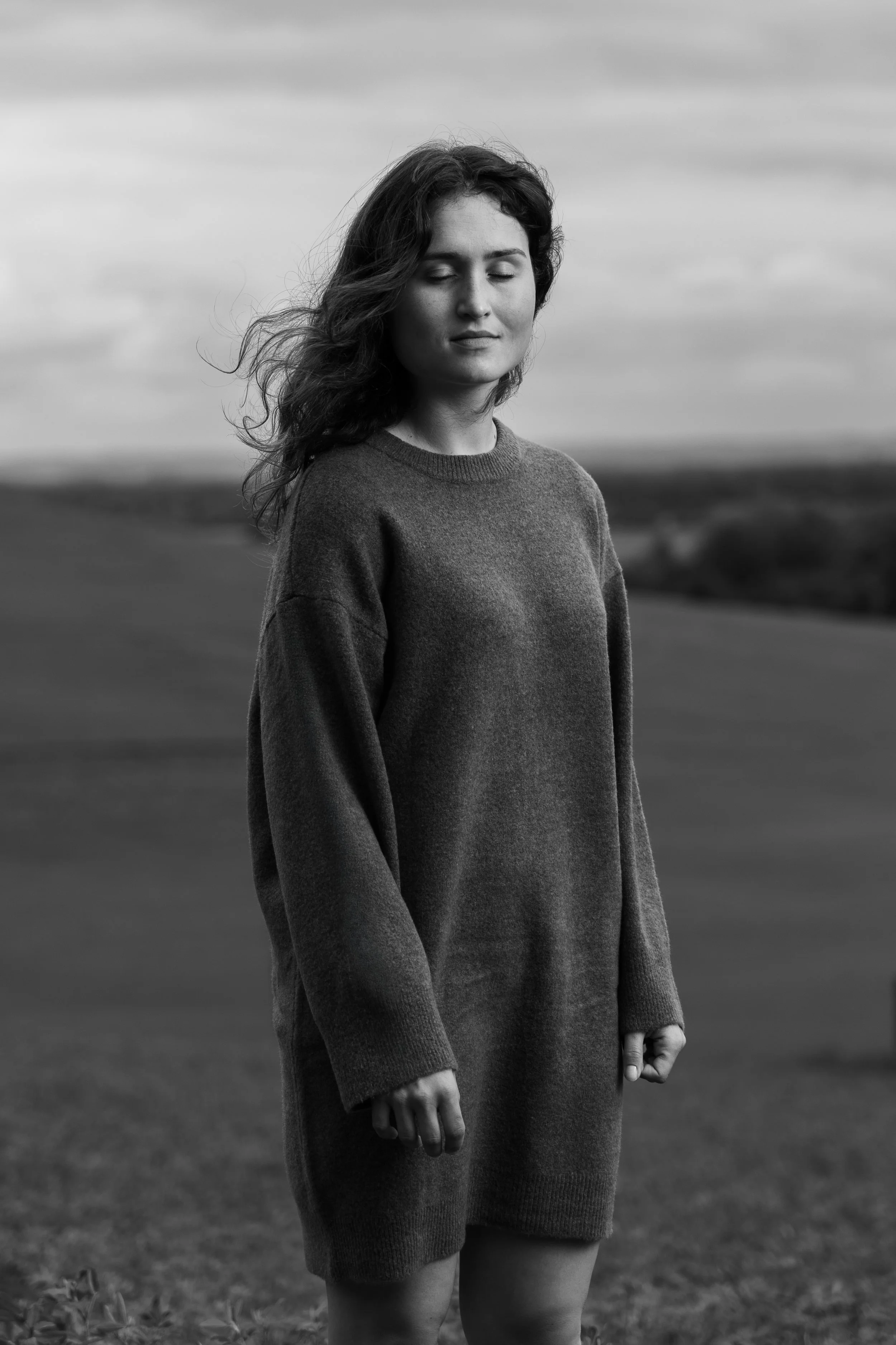Black-and-white photo of a woman standing outdoors with eyes closed, wearing a long-sleeved sweater dress, with a blurred landscape in the background.