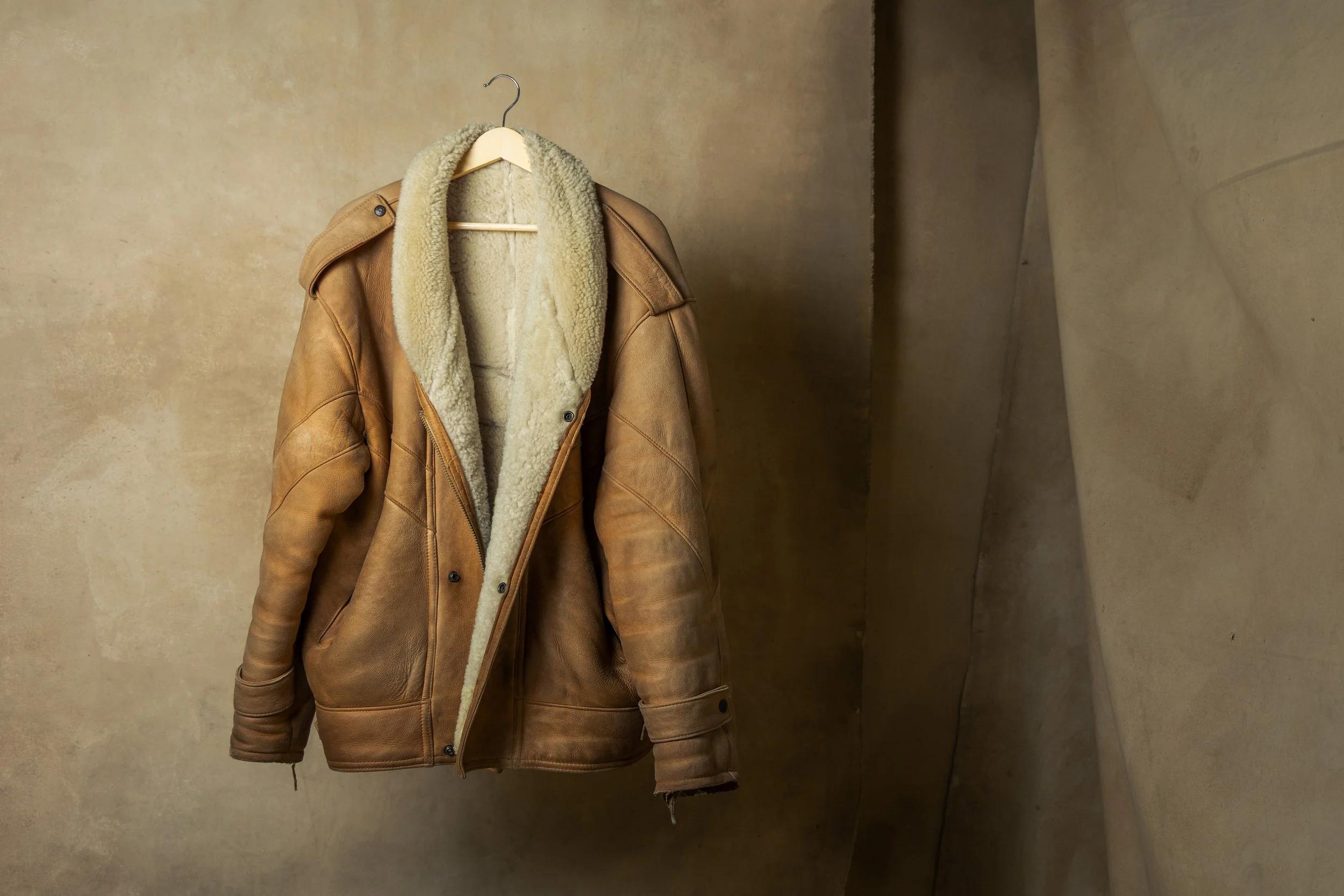 Brown leather shearling jacket hanging on a wooden hanger against a beige wall.