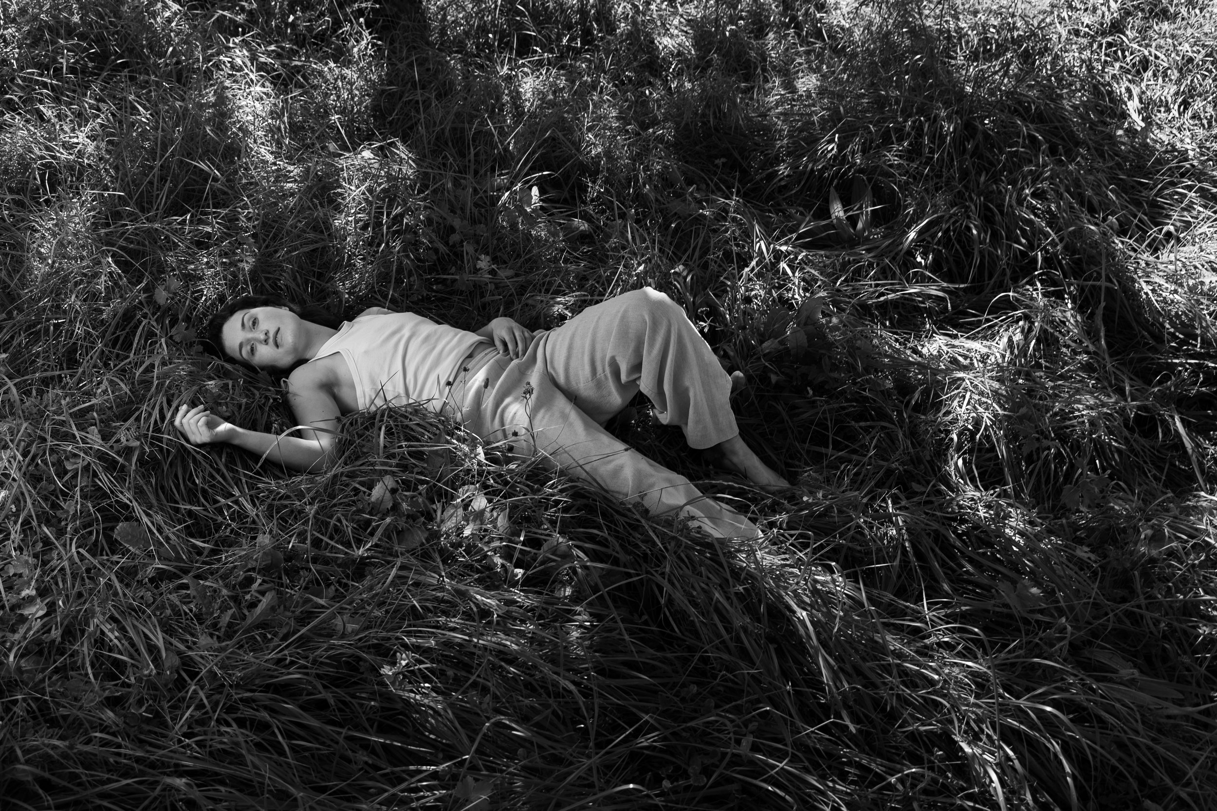 A woman lying on the grass in a natural outdoor setting, wearing a sleeveless top and loose pants, with sunlight creating shadows around her.