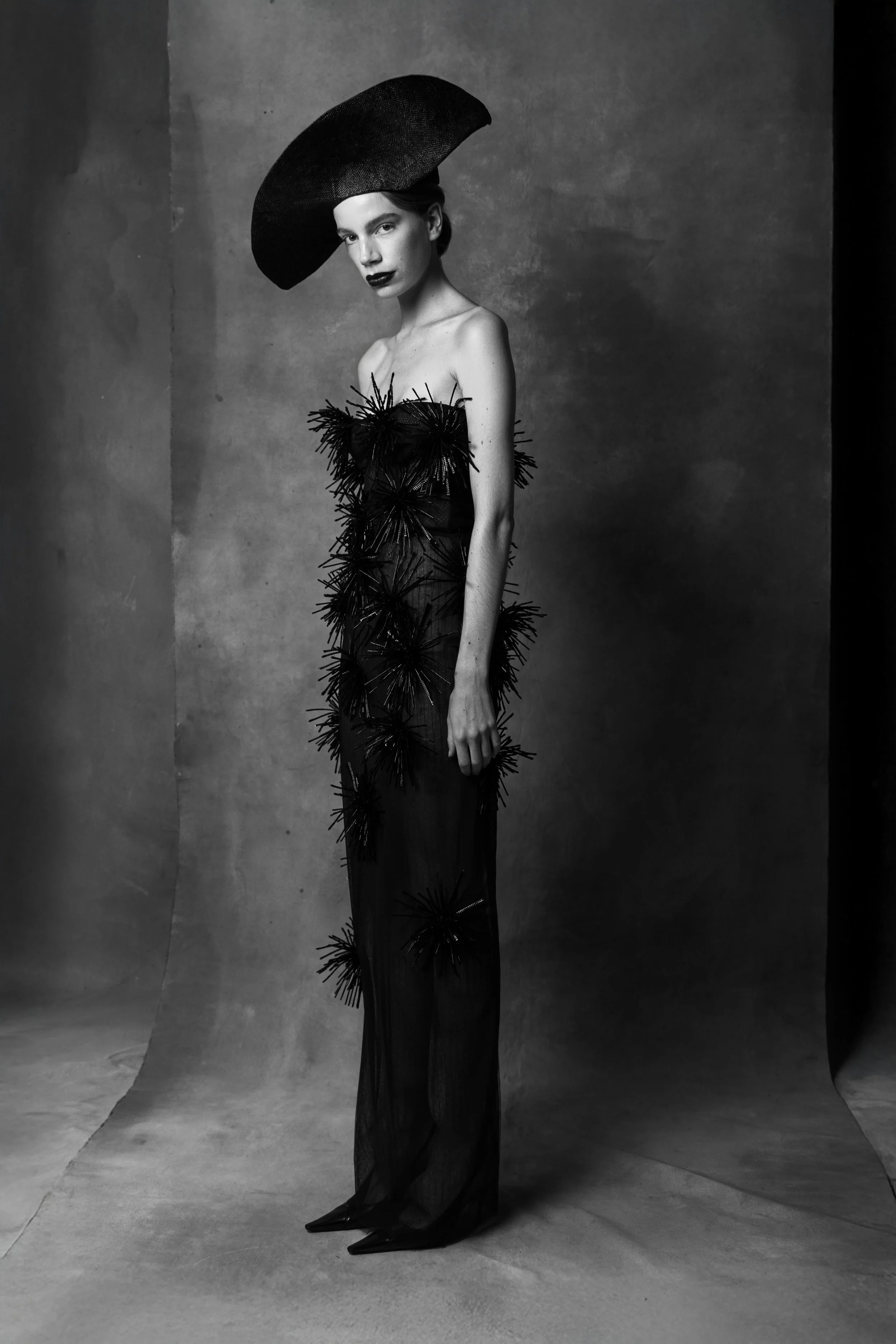 A woman in a black dress with textured, spiked embellishments stands against a plain backdrop, wearing a large black hat and dark lipstick, in a black and white photo.