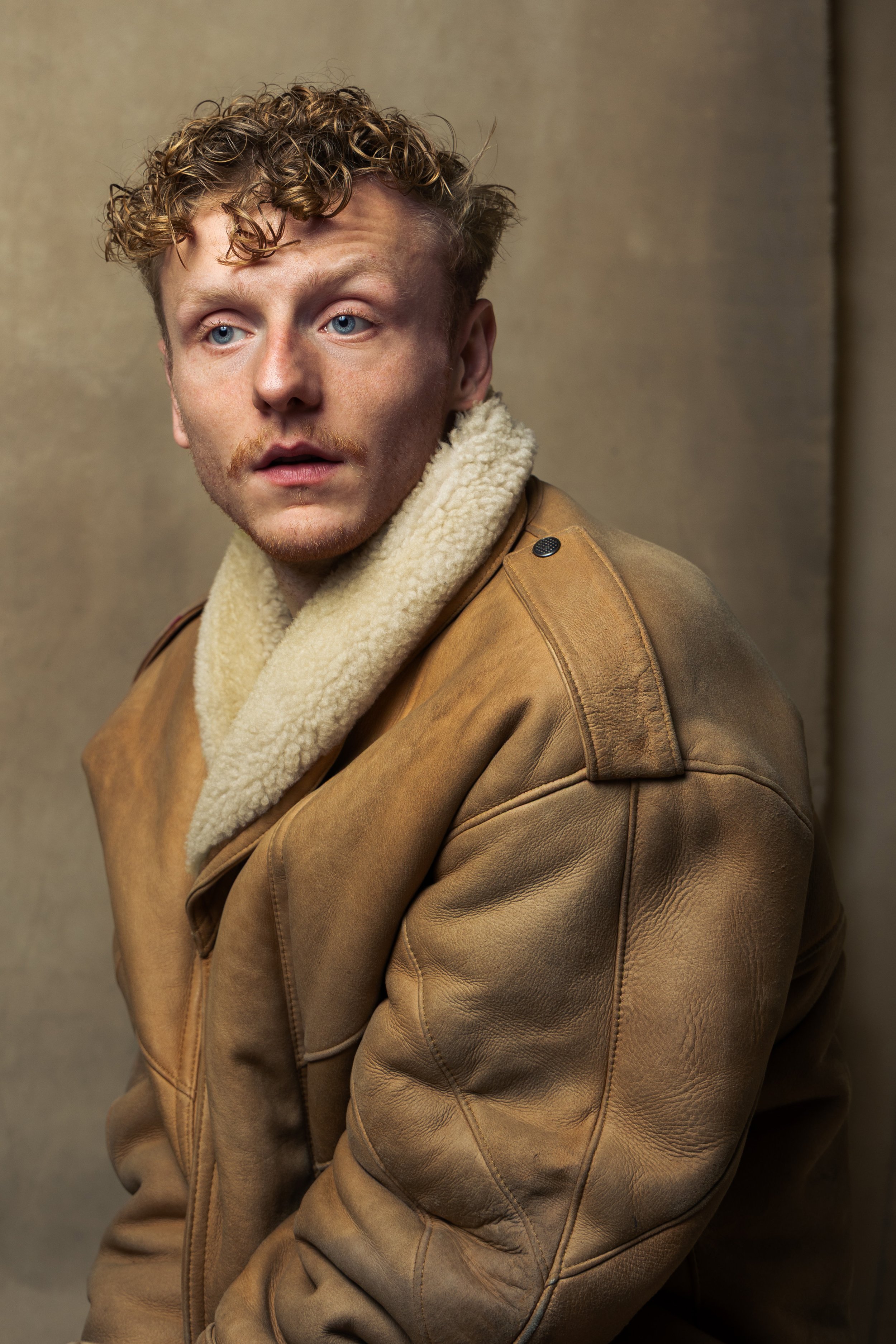 A young man with curly blonde hair and blue eyes wearing a tan leather jacket with a shearling collar, sitting against a plain brown background.