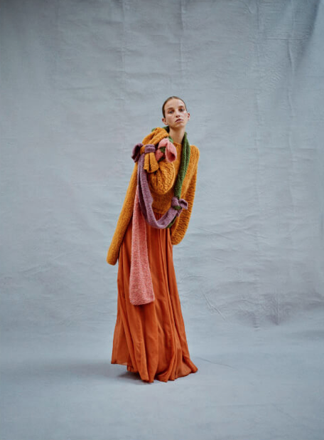 A woman standing in front of a plain gray background, wearing a colorful winter outfit that includes a mustard yellow coat, a long orange skirt, and a multicolored scarf with pink, purple, and yellow sections.