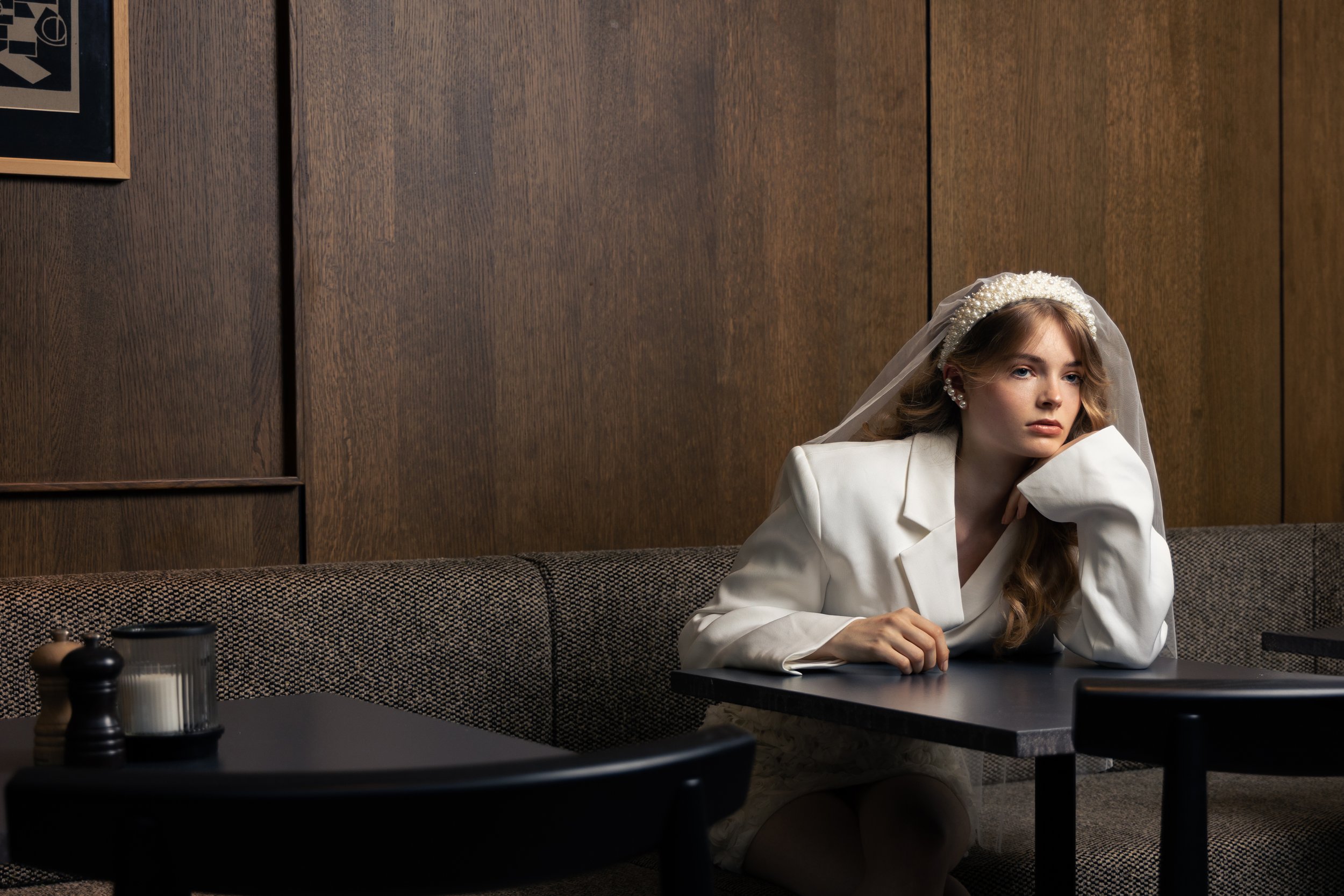 A young woman wearing a white blazer and floral bridal headband with a veil, sitting alone at a dark table in a restaurant or cafe, resting her head on her hand, looking thoughtful.