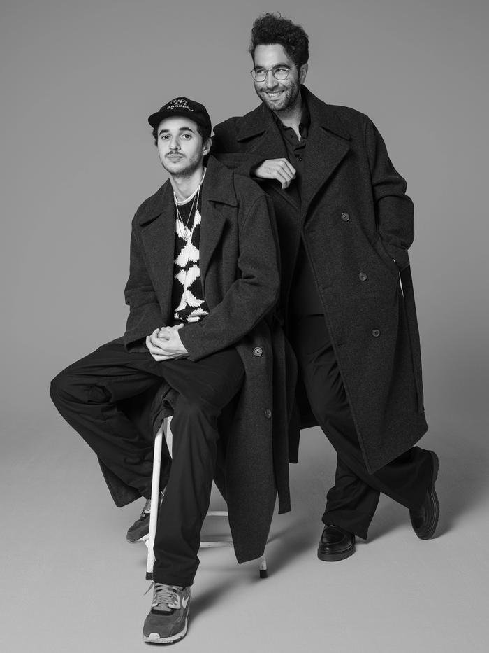 Two men in dark coats posing together against a plain background, one sitting on a stool and the other standing with a hand on his shoulder, both smiling and looking at the camera.