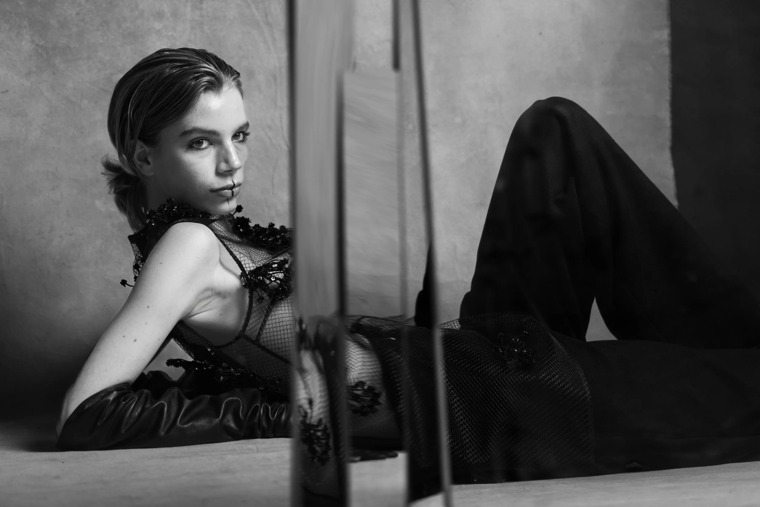 A black and white photo of a woman lying on the floor, wearing a lace top and gloves, with a mirror partially obscuring her lower body.