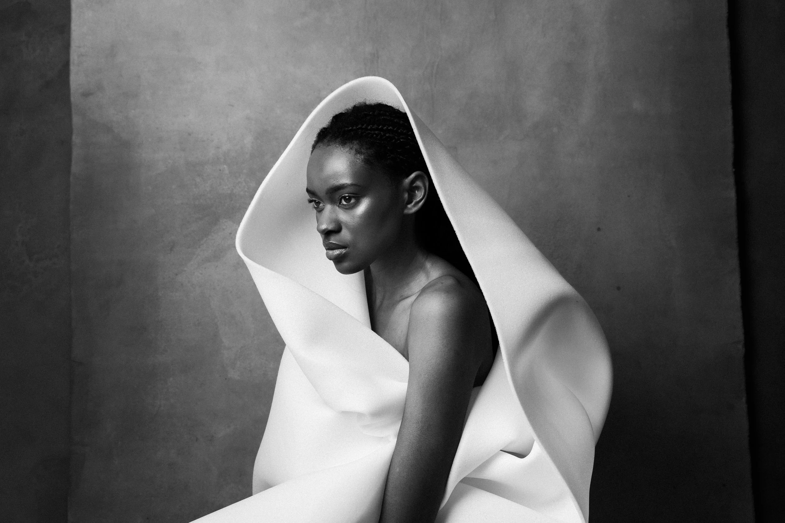 A black-and-white photograph of a woman with braided hair, looking away from the camera, draped in an artfully arranged high-fashion white fabric around her shoulders and body.