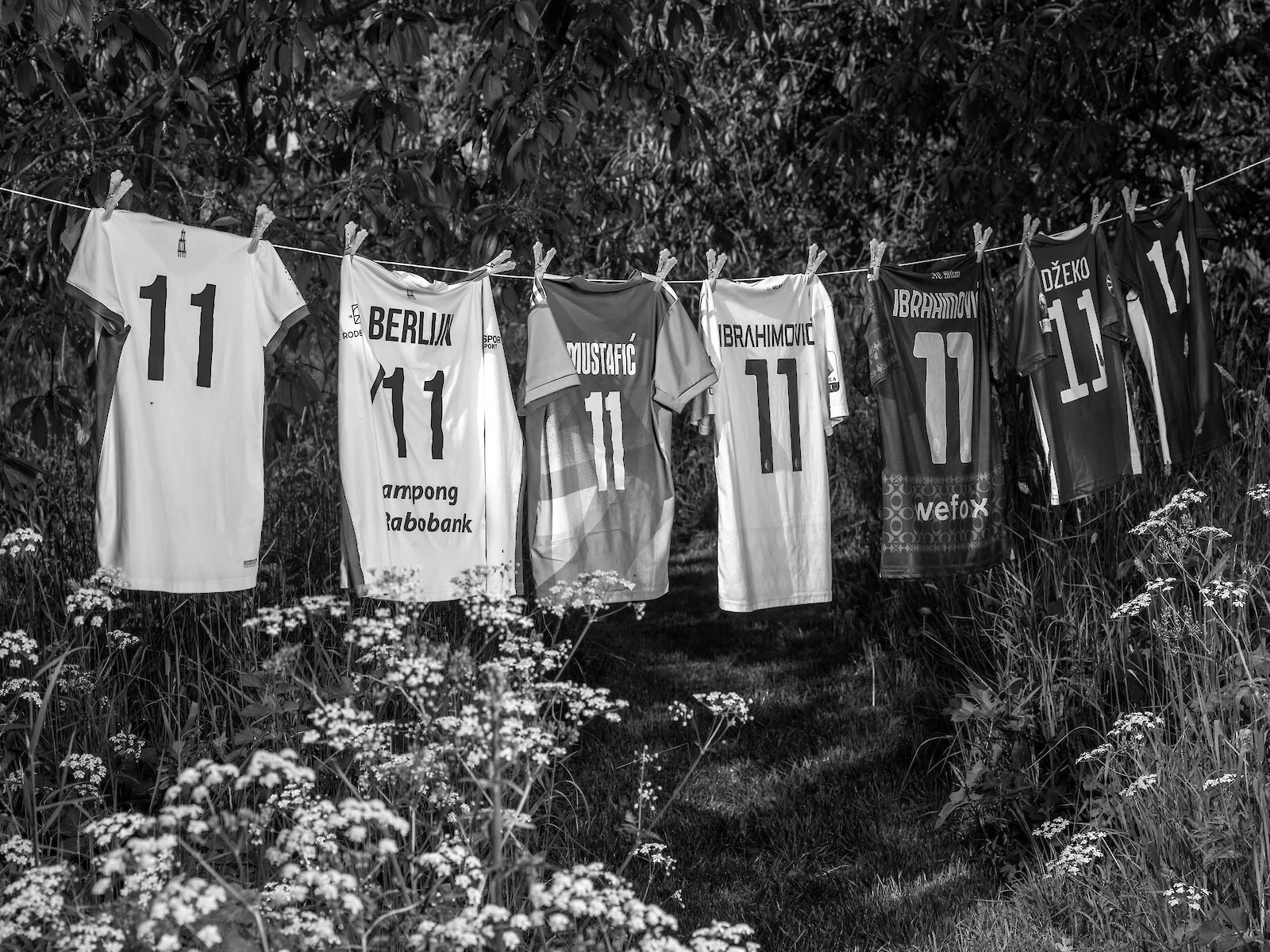 Multiple soccer jerseys hanging on a line amidst grass and wildflowers, with some jerseys displaying the number 11 and various player names.