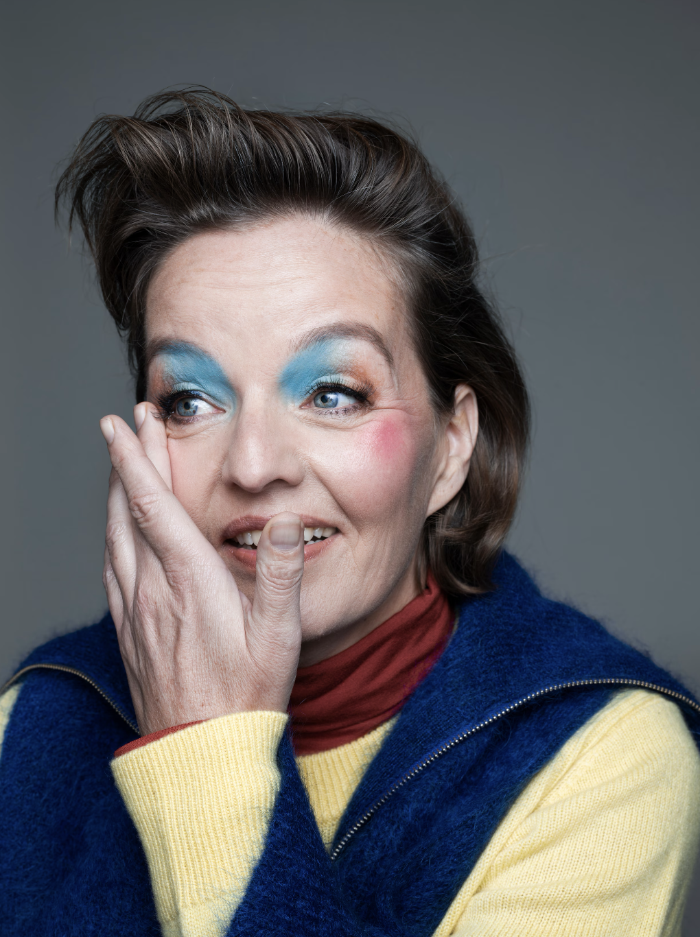 A woman with blue, orange, and gold eyeshadow, pink blush, and a red lipstick, wearing a blue jacket over a yellow sweater with a red turtleneck, touching her face with her left hand.