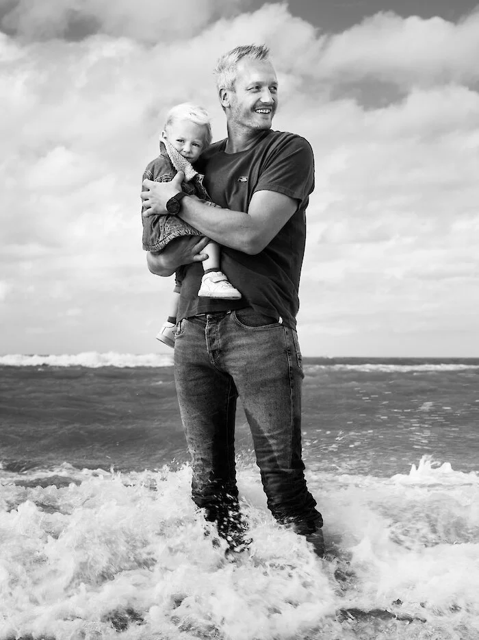A smiling man holding a young girl in his arms while standing in the ocean with waves around their feet, at the beach.