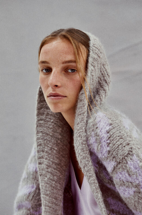 Young woman wearing a gray, fuzzy knit hood and matching cardigan, looking at the camera with a neutral expression against a plain background.