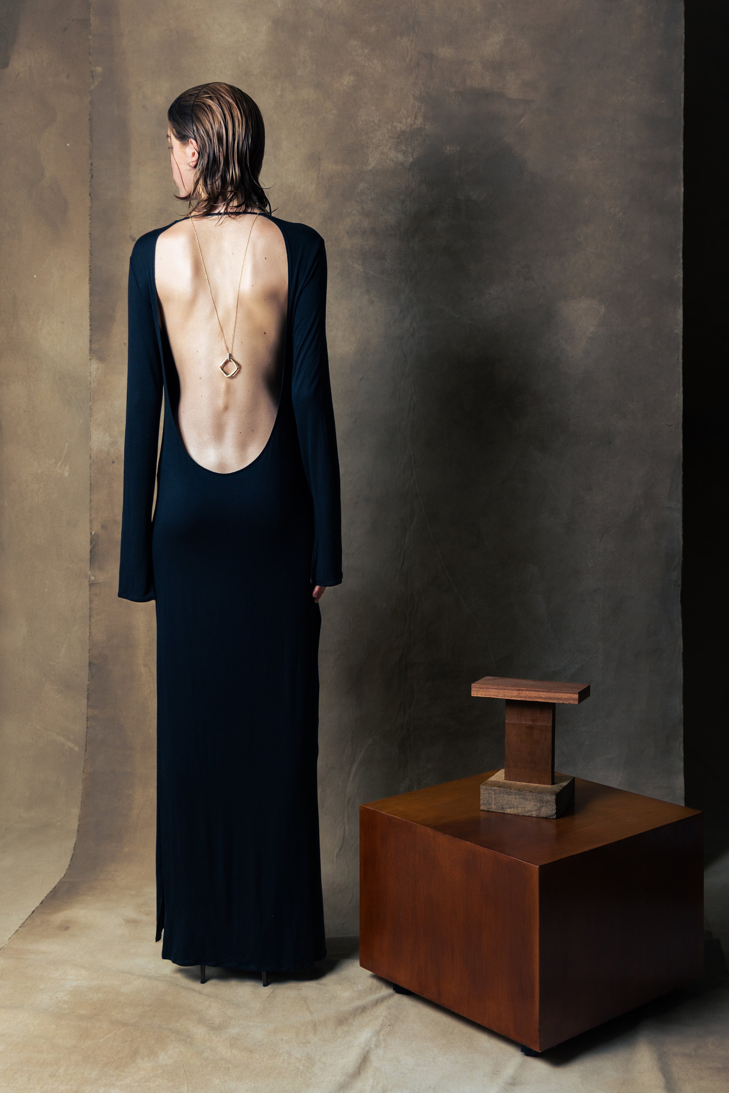 A woman with shoulder-length hair is dressed in a long, elegant black dress with an open back, standing next to a wooden table with a small modern sculpture on it, against a neutral textured wall.