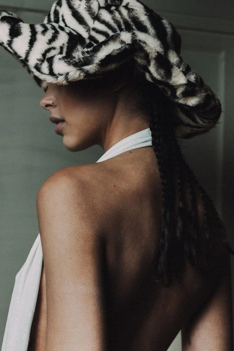 A woman wearing a large, zebra-patterned hat and a white scarf around her neck, with long braided hair.