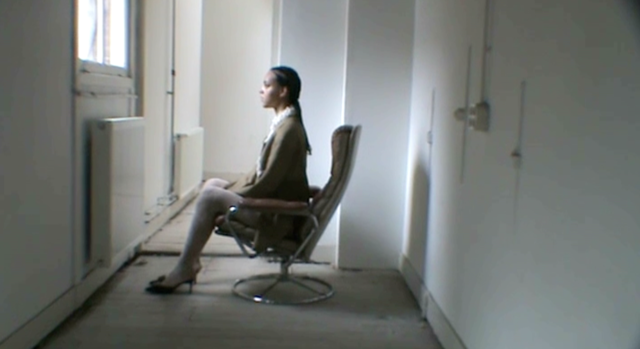 A woman sitting on an office chair in a dimly lit room, facing a window with her eyes closed.