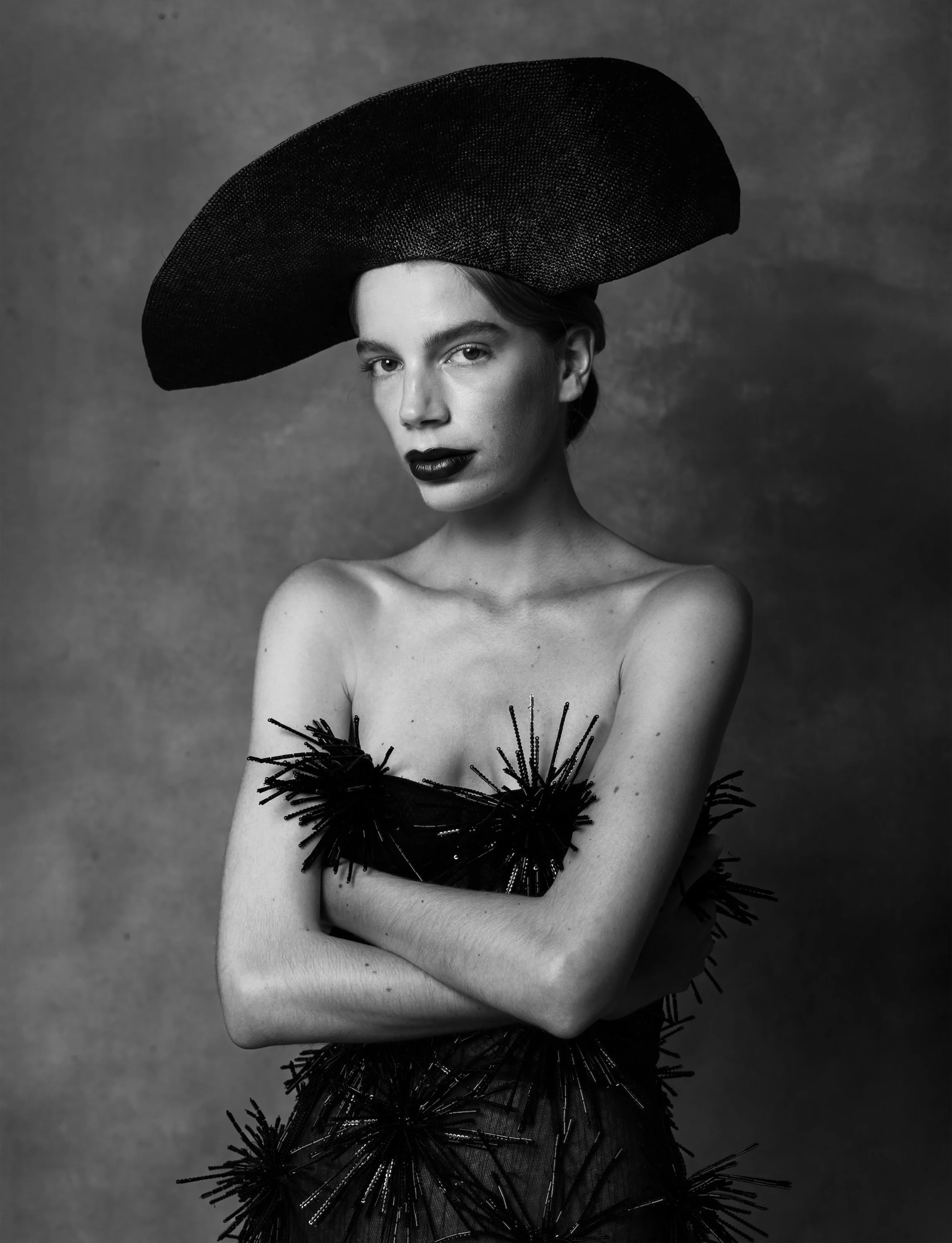 Black and white portrait of a woman wearing a large, wide-brimmed hat and a dress with embellishments, posing with arms crossed in front of her chest.
