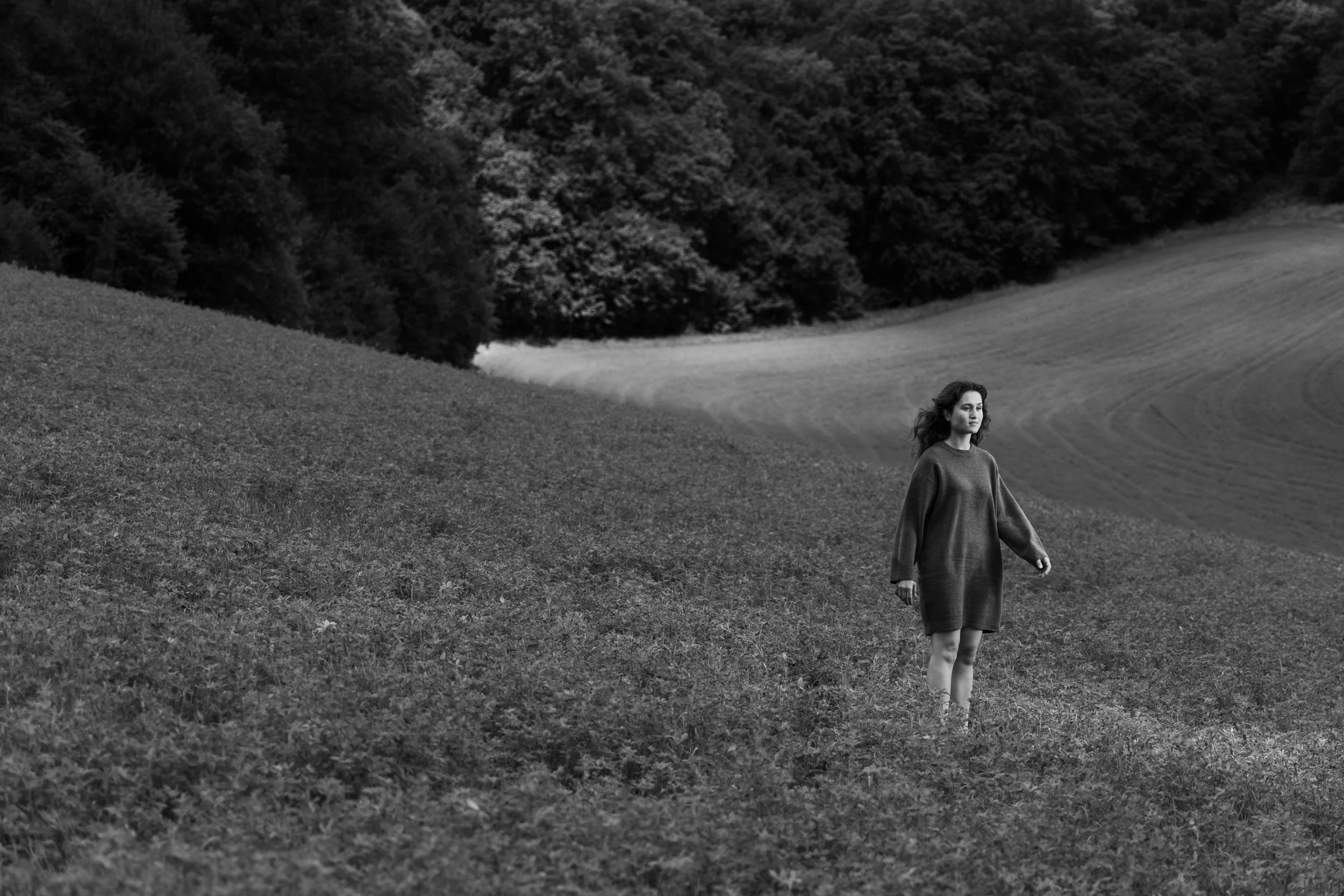A woman in a oversized sweater dress standing alone on a grassy hillside with a forest and rolling fields in the background, all in black and white.