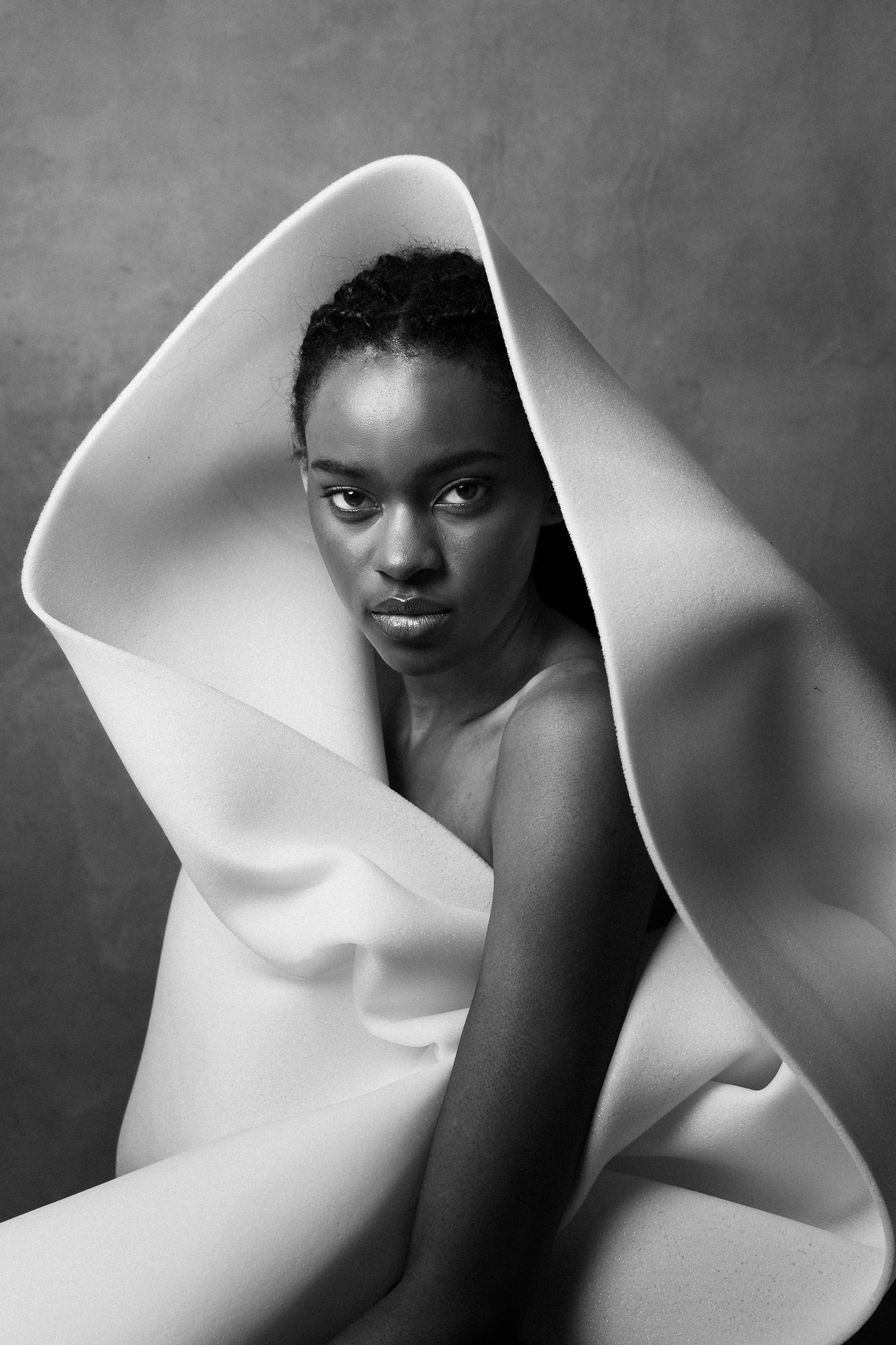 A black and white photo of a woman with dark skin, looking directly at the camera, partly wrapped in a large piece of fabric or material.