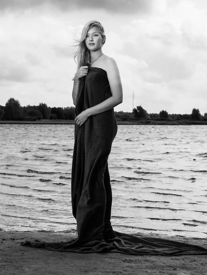 A woman standing on the shore near water, wrapped in a dark towel, with wind blowing her hair, in black and white.