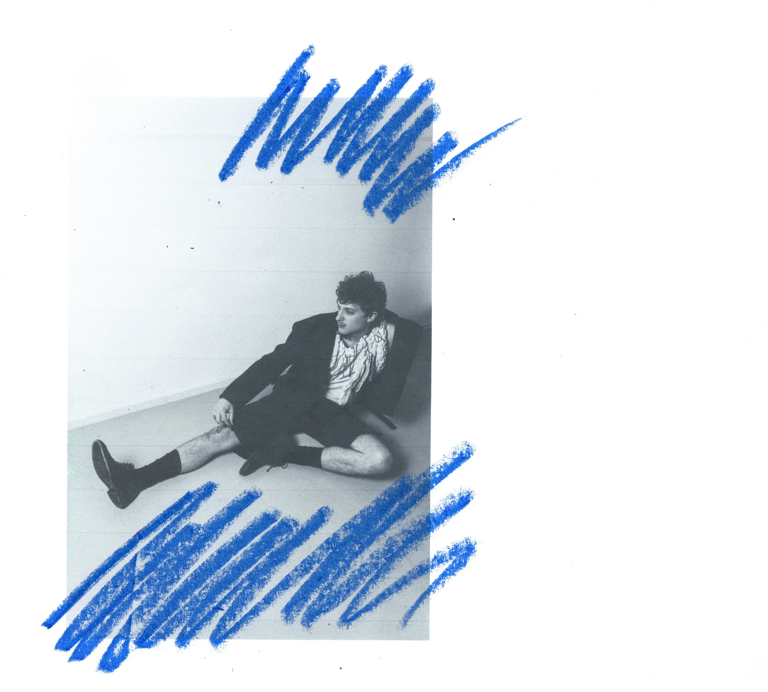 A black-and-white photo of a young man sitting on the floor, wearing a blazer, shorts, and sneakers, with scribbled blue markings surrounding him.