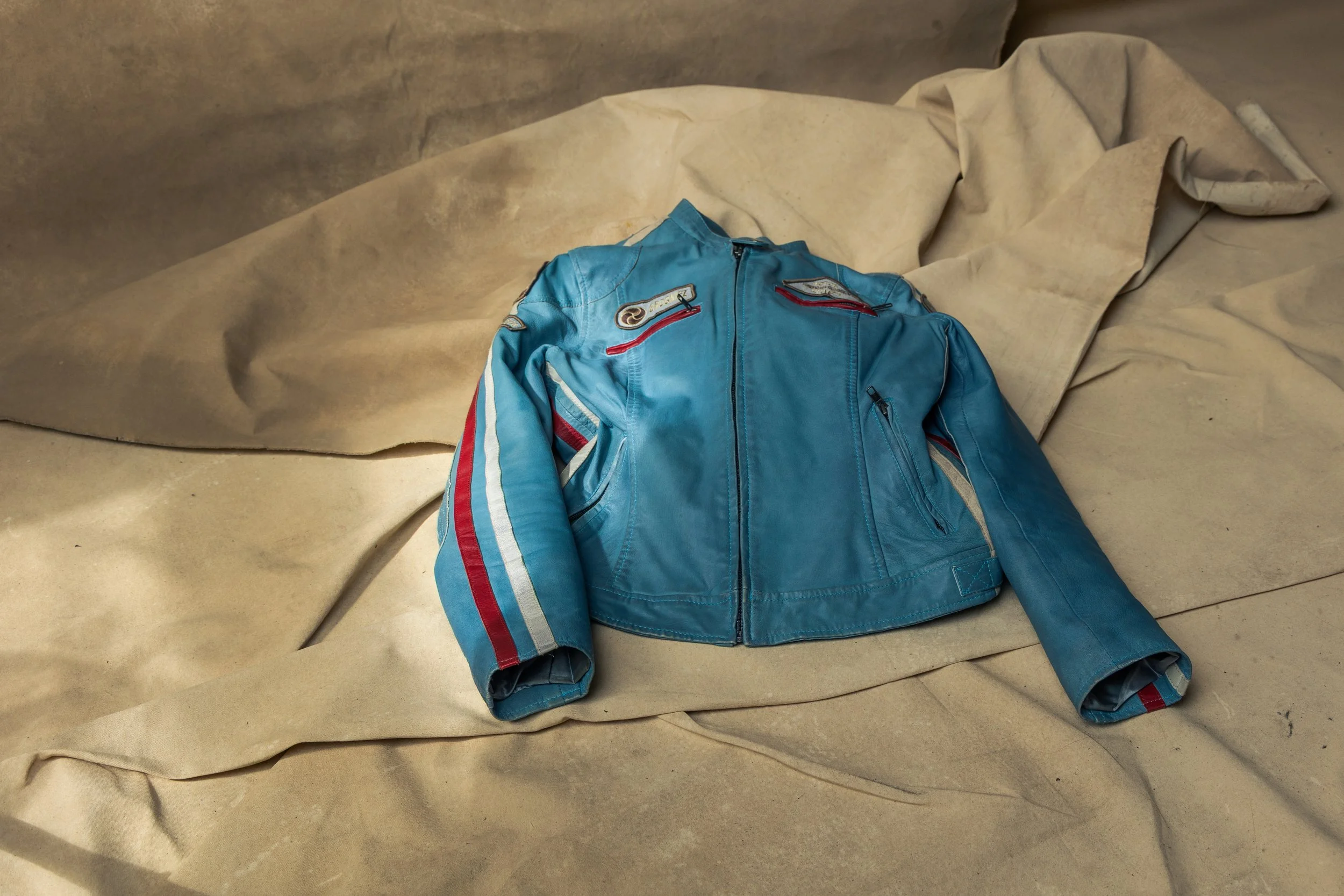 A blue leather jacket with red, white, and gray stripe details, lying on beige crumpled paper or fabric.
