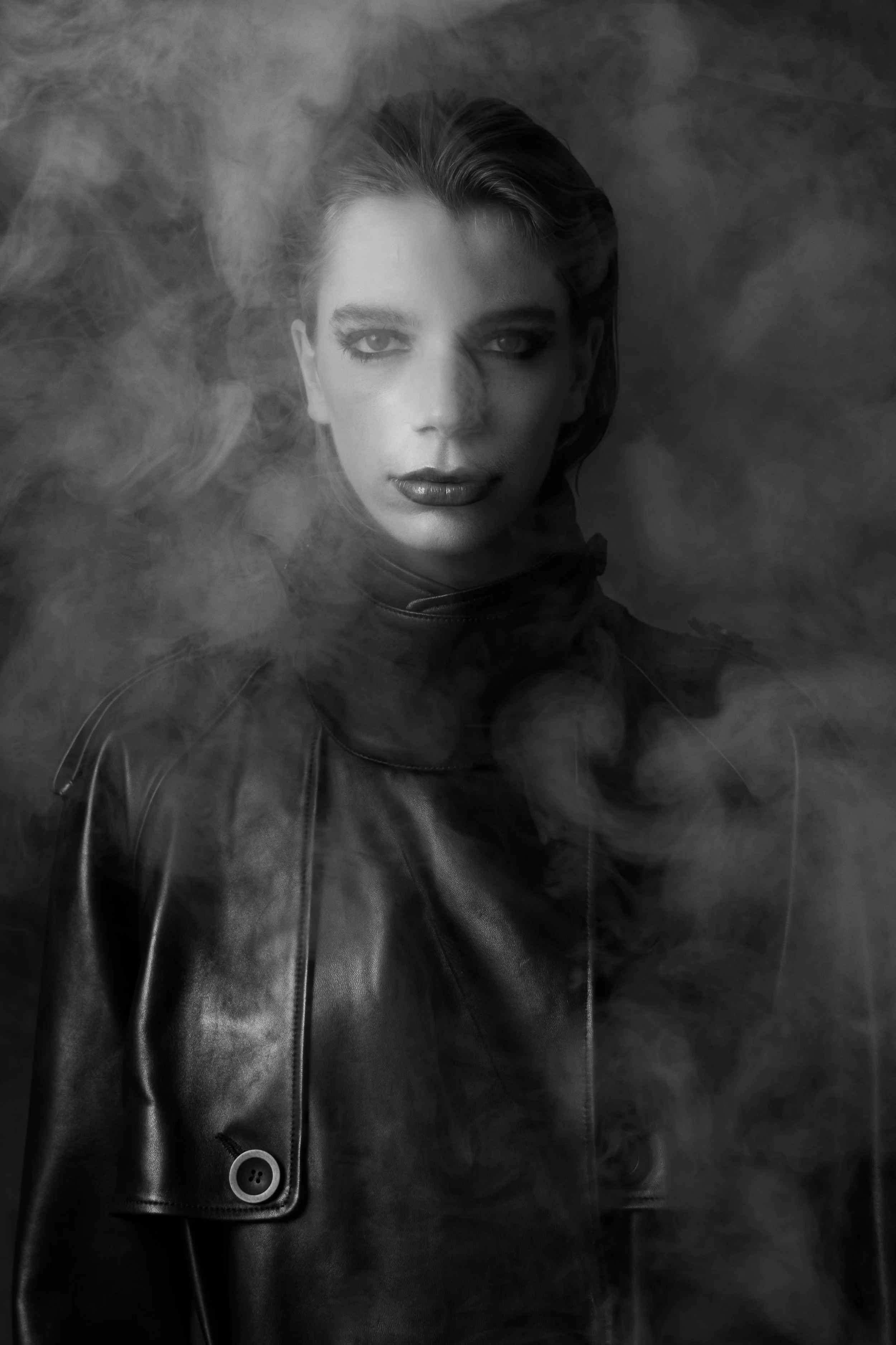 Black and white portrait of a woman wearing a leather jacket, with smoke surrounding her, and striking makeup with dark lipstick and eye makeup.