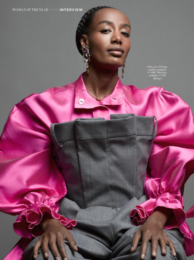 Woman with braided hair and elegant earrings, wearing a shiny pink blouse with voluminous sleeves and a grey wrap dress, sitting against a grey background.