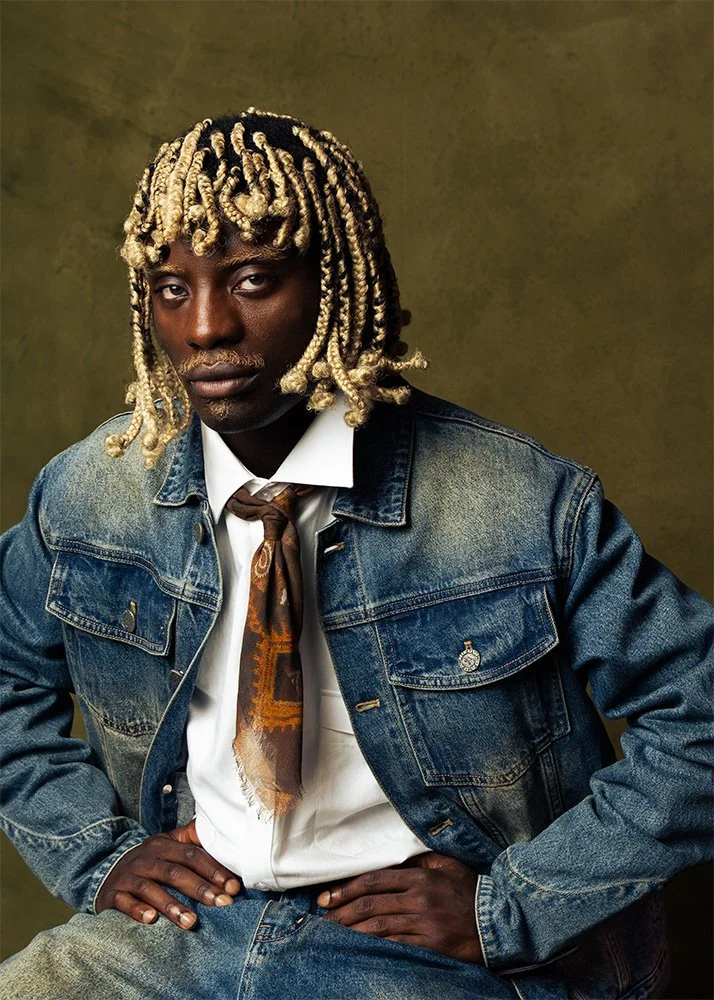 A man with dreadlocks and dark skin wearing a denim jacket, white shirt, necktie, and sitting with hands on his hips against a plain background.