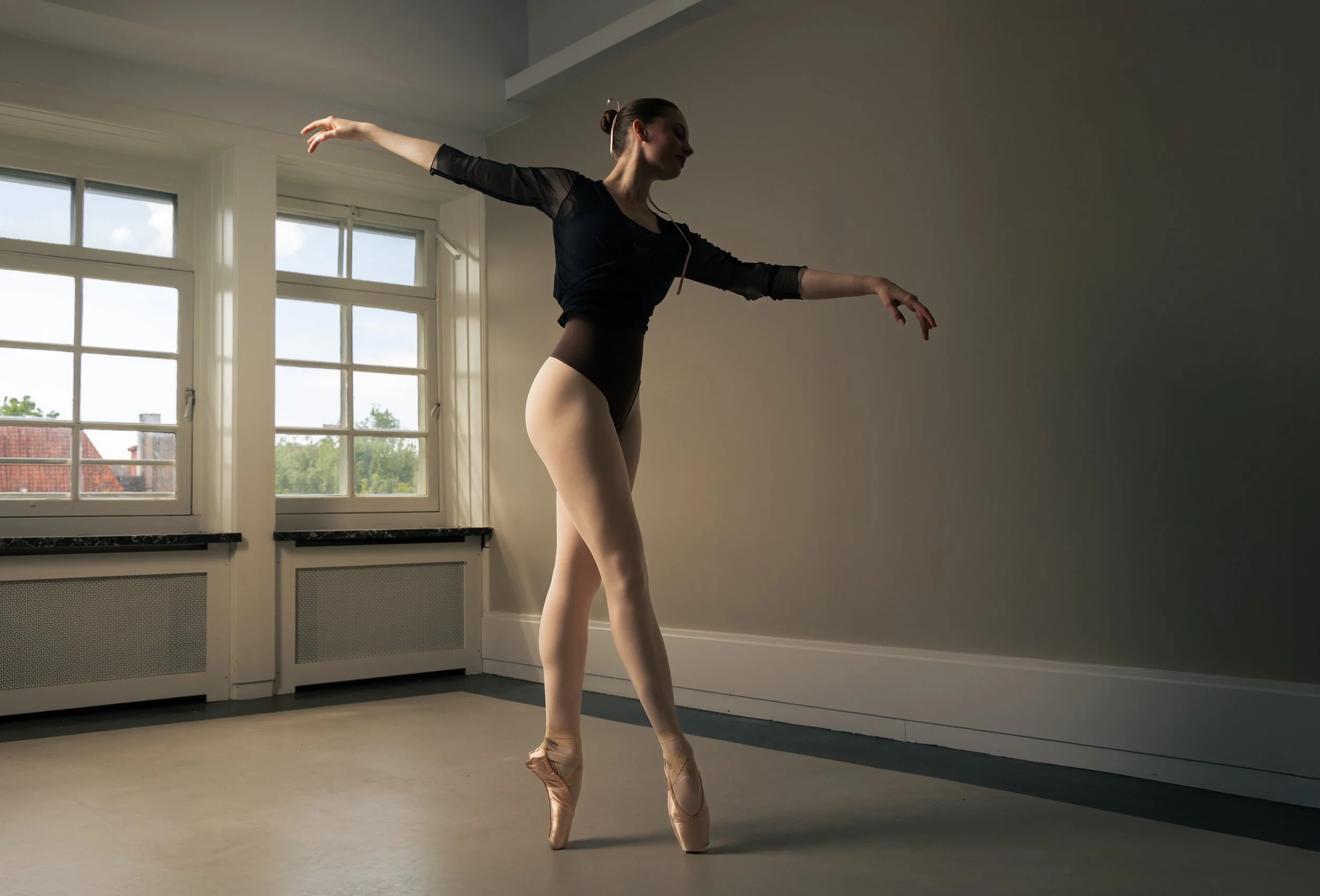 Ballet dancer practicing in a classroom, standing en pointe with arms outstretched, near large windows.