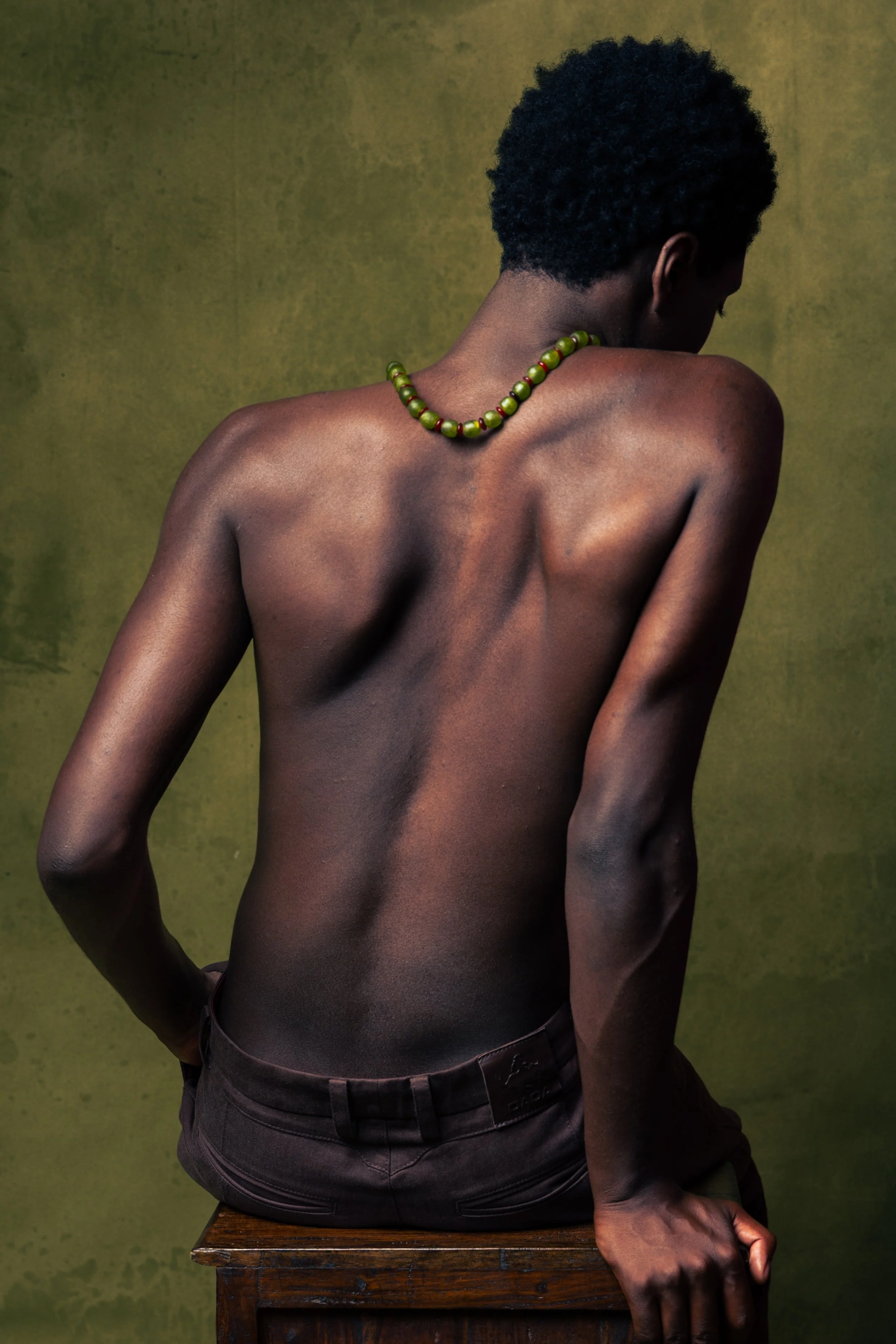 Back view of a shirtless person with dark skin and short curly hair, wearing a green beaded necklace, sitting on a wooden stool against a textured green background.