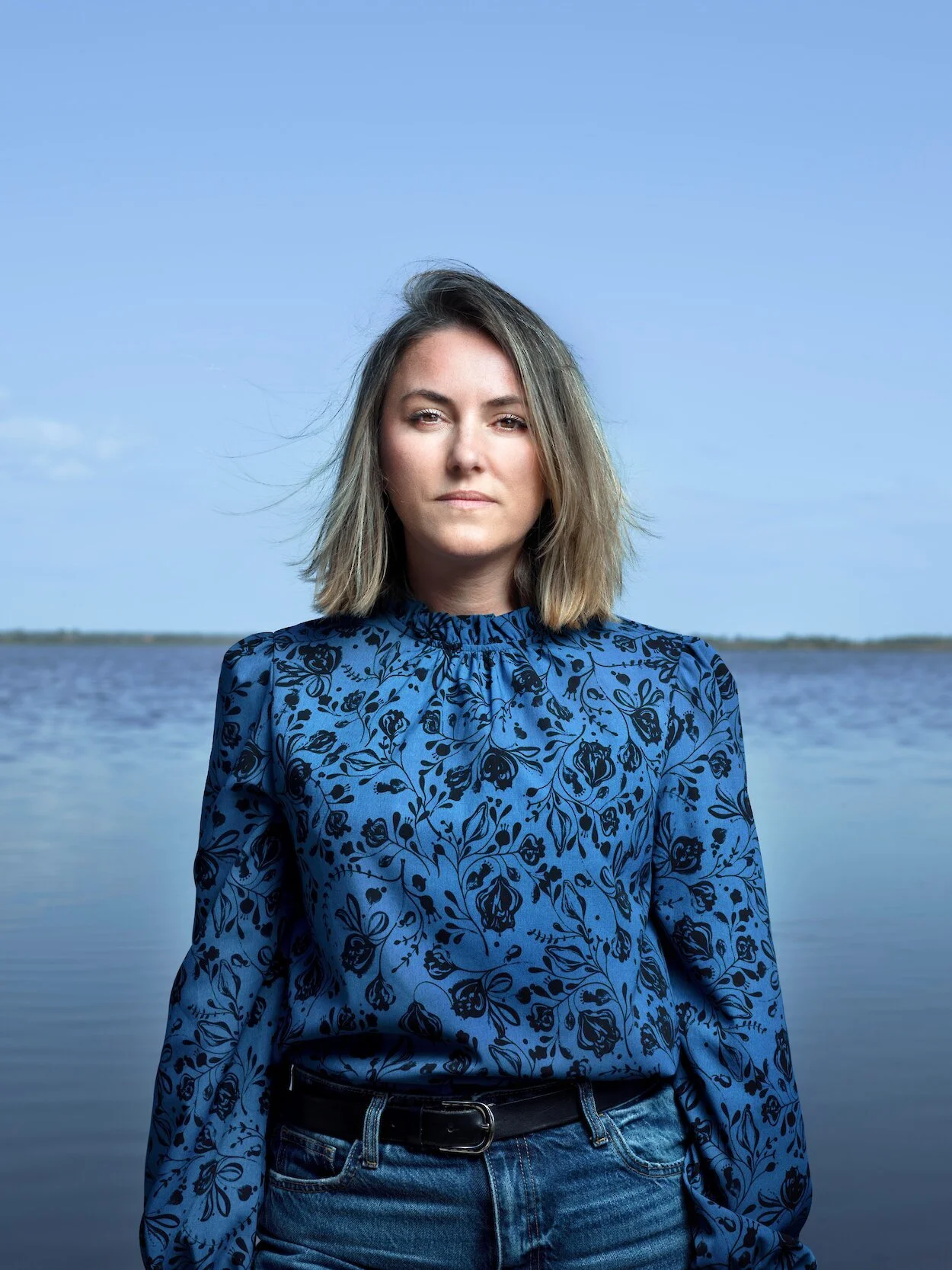 A woman with shoulder-length blonde hair stands outdoors by a body of water under a blue sky, wearing a blue floral long-sleeve blouse and jeans.