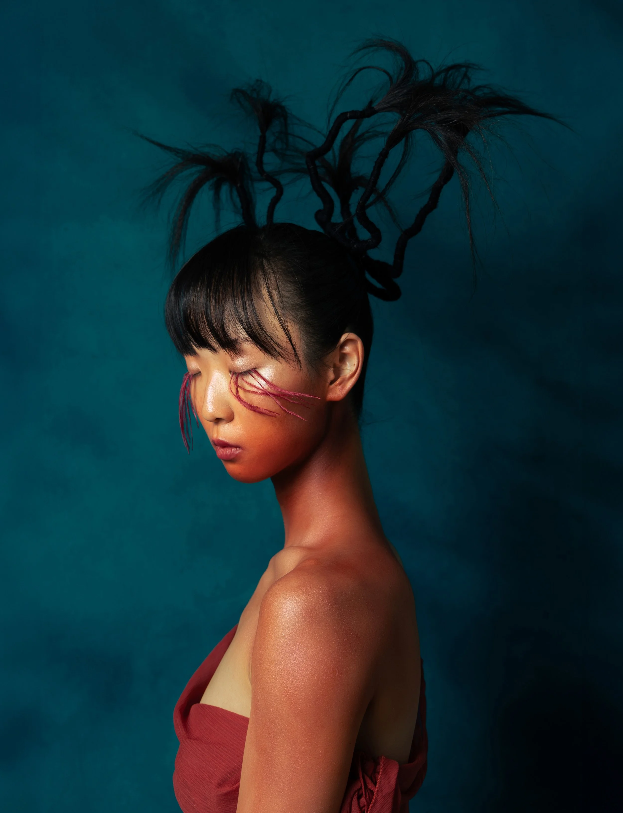 Artistic portrait of a woman with short black hair, eyes closed, and face turned aside. She has pink and red painted lines on her face, and her hair is styled in upwards, twisted dreadlocks. She is wearing a red off-shoulder top, against a dark teal background.