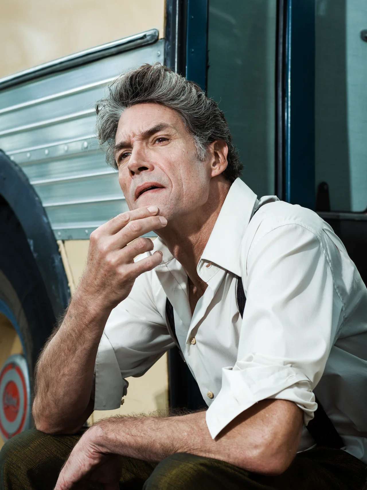A middle-aged man with gray hair and a white shirt, appearing contemplative or worried, sitting with his elbow on his knee and hand near his chin outdoors, with a trailer or mobile home behind him.