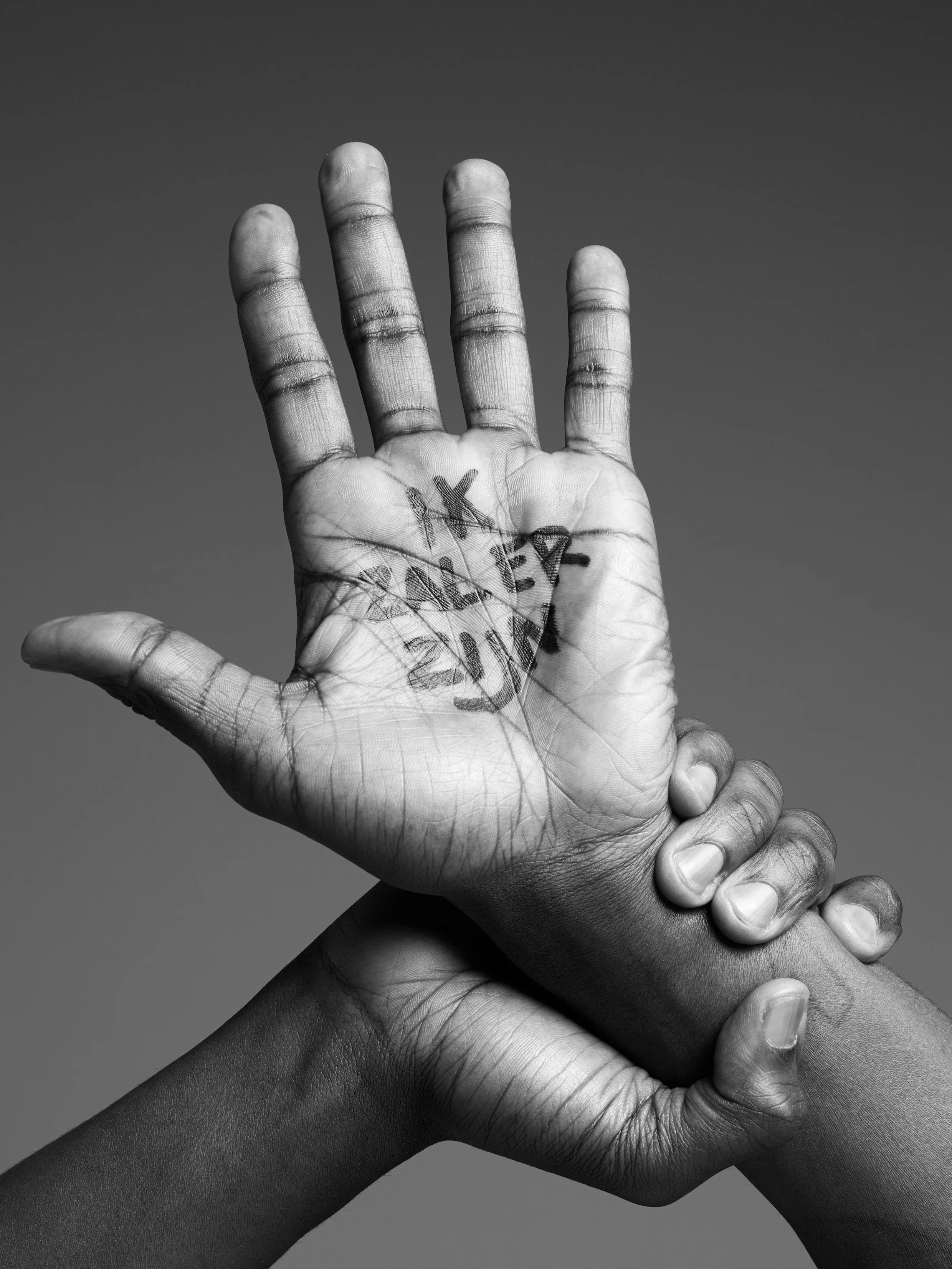 Black and white photo of two hands clasped, with one hand raised showing the palm. The palm has the words 'IK ZAL ER ZIJN' written on it. The background is plain and dark.