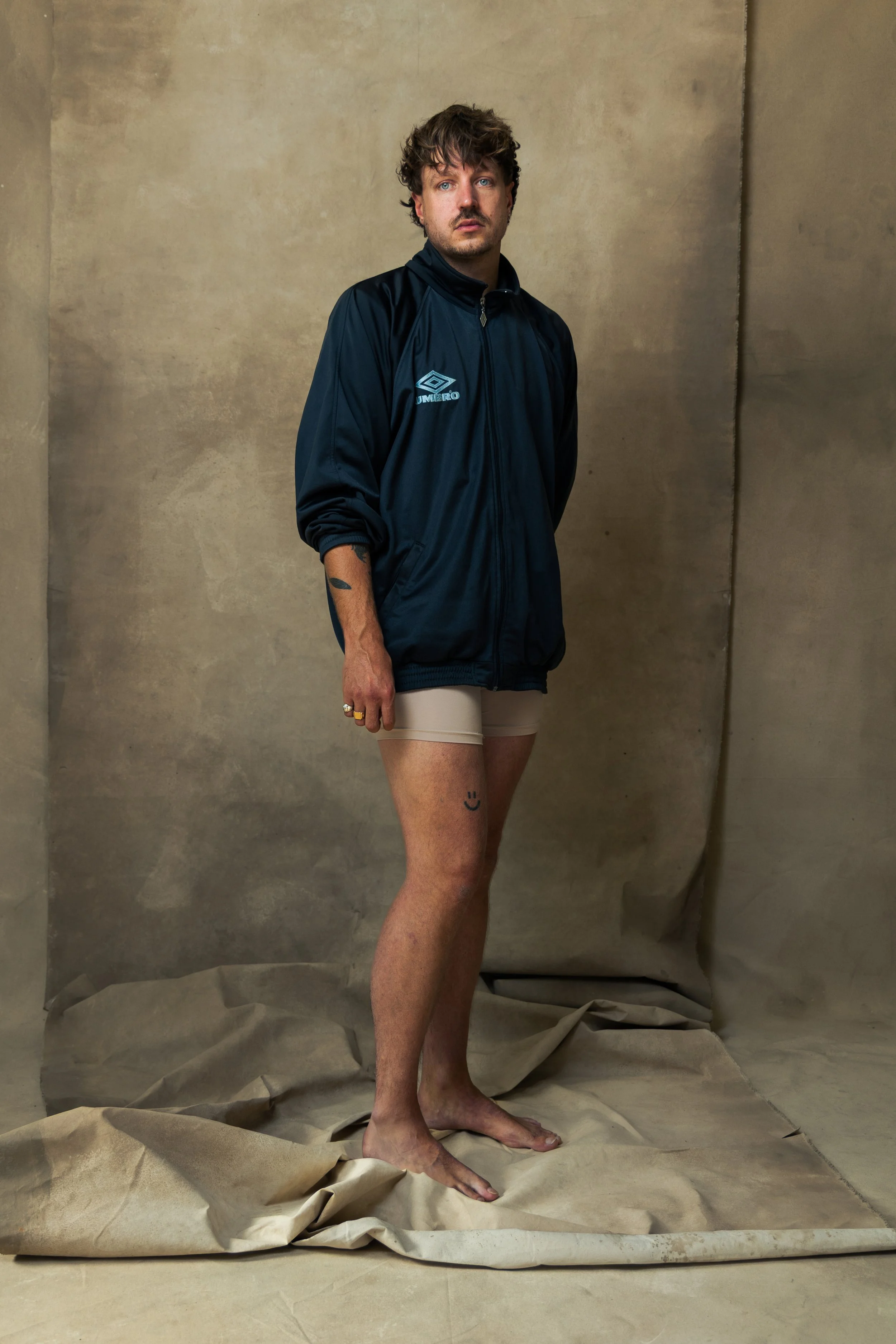 A man with curly hair and a beard standing barefoot on a crumpled paper backdrop, wearing a dark sports jacket and beige shorts, with a small smiley face tattoo on his right thigh.