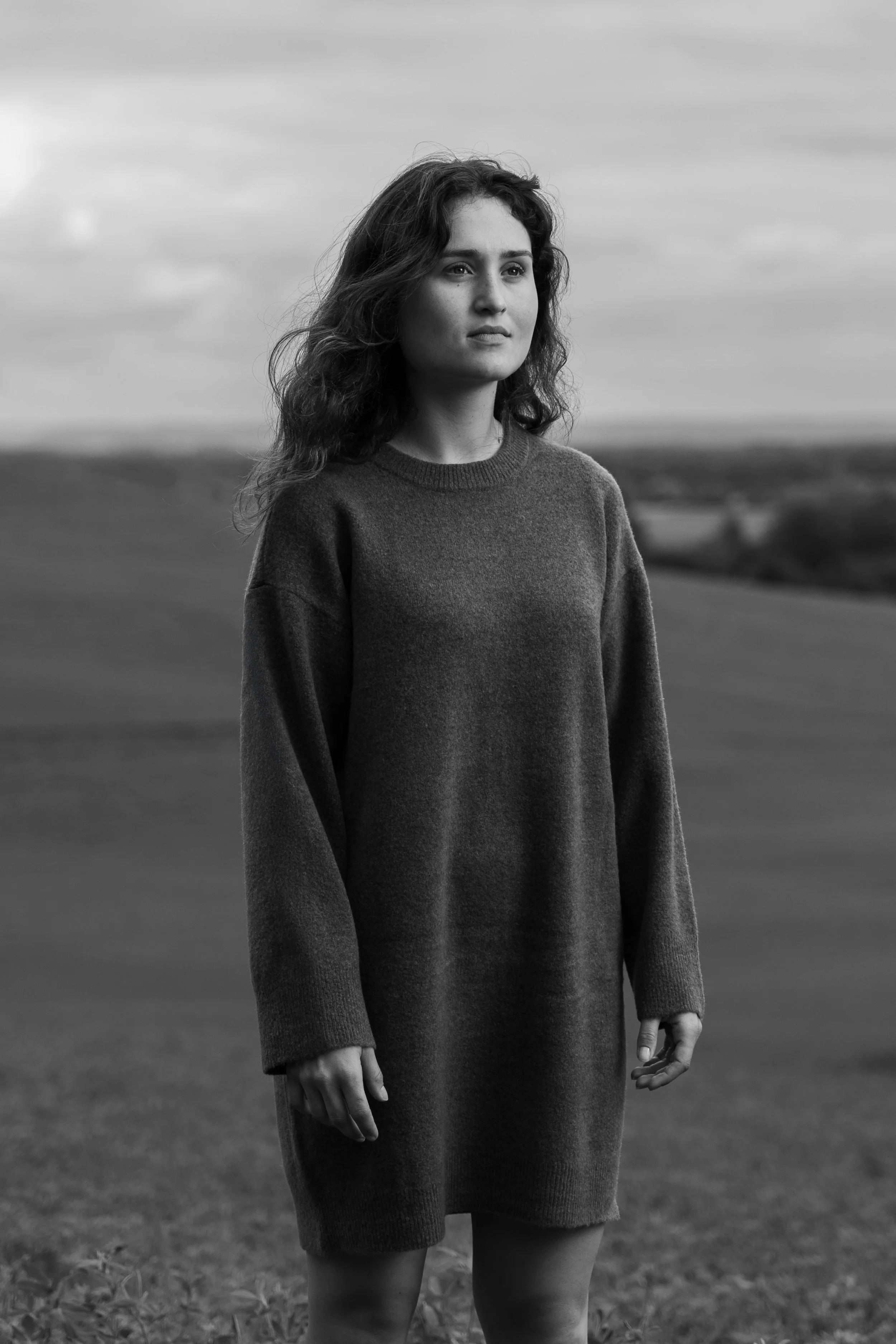 A woman stands outdoors in a field, looking into the distance with a serious expression, wearing a long sweater dress. The photo is in black and white.