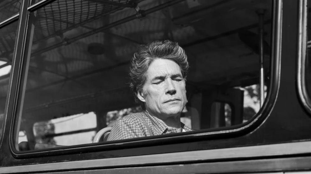 A man with closed eyes and a serious expression sitting in a vehicle, viewed through a window.