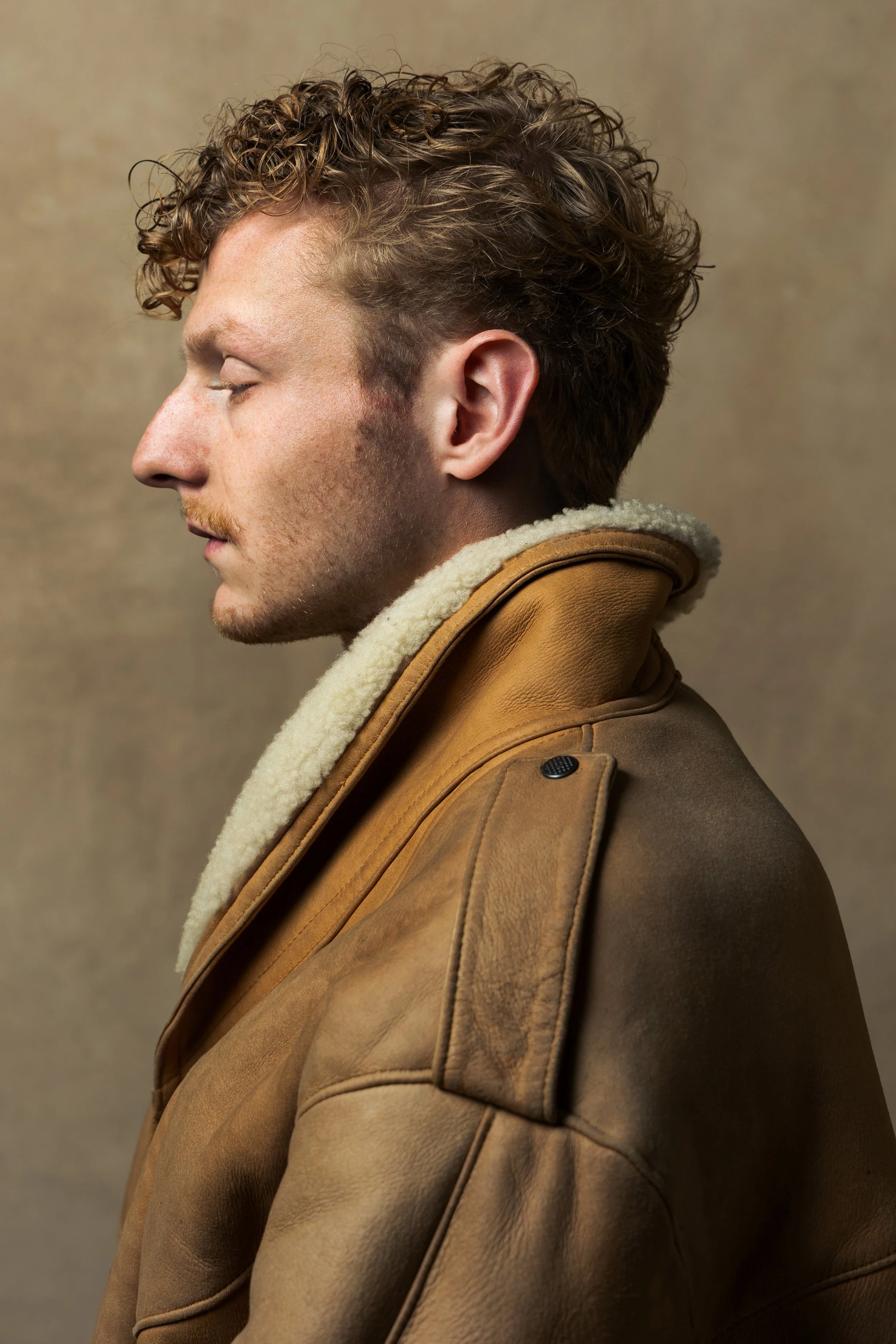 A side profile of a young man with curly hair and a light beard, wearing a brown leather jacket with a shearling collar, against a neutral background.
