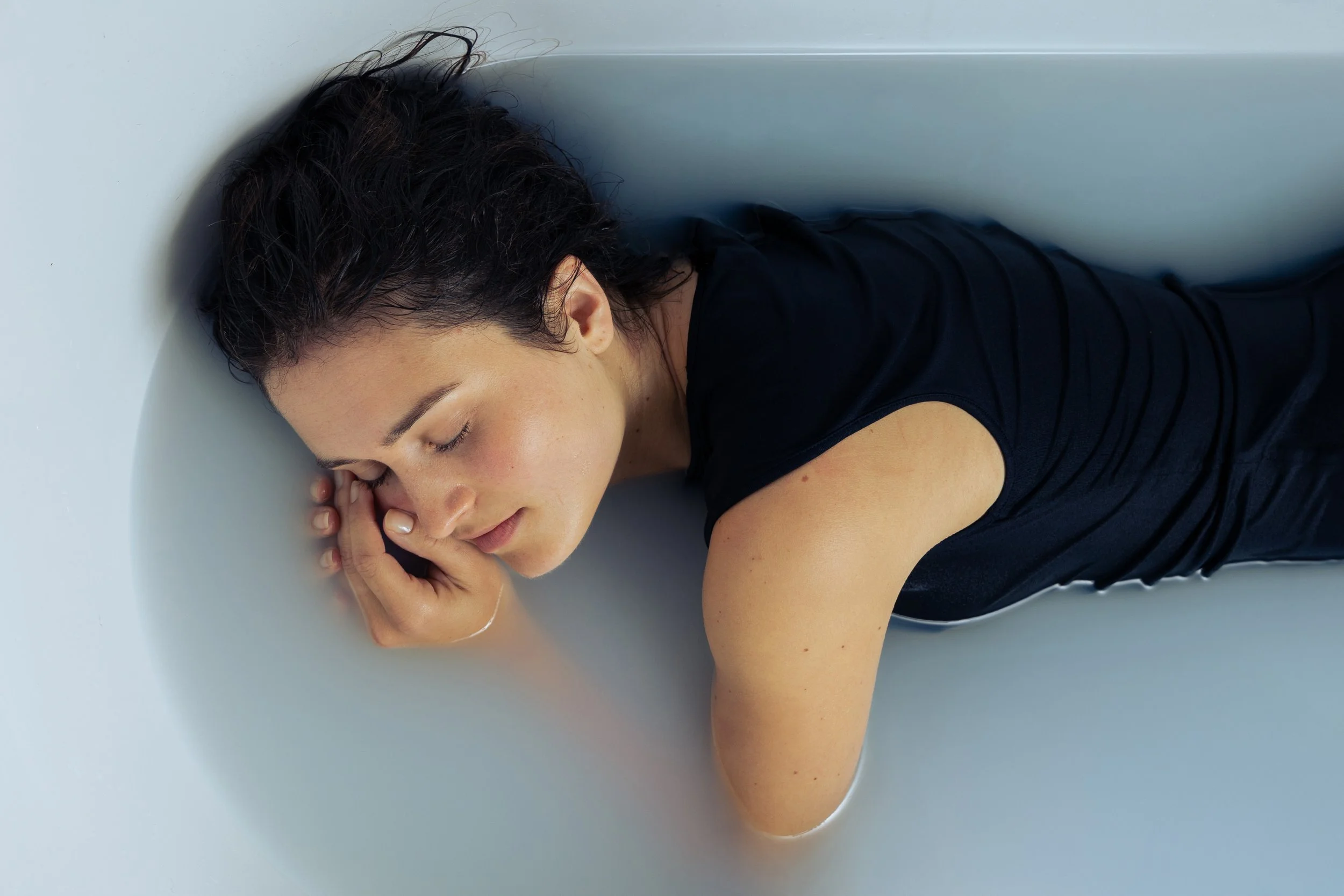 A woman with dark hair lying on her side in a bathtub filled with milky white water, wearing a black sleeveless top, with her eyes closed and a peaceful expression.