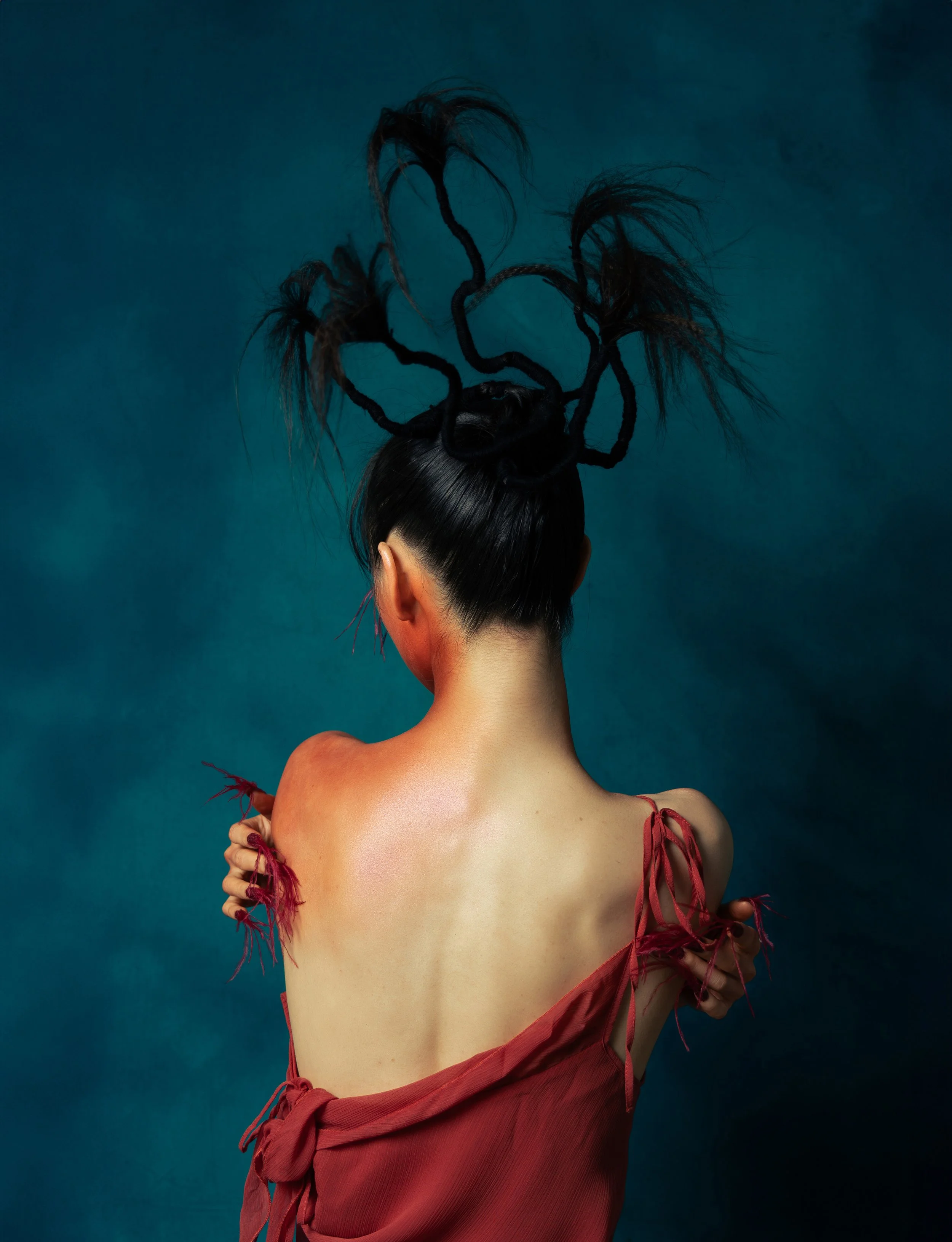 A woman with dark hair styled in an elaborate fashion with twisted, teased, and stick-straight sections, wearing a red off-shoulder dress with torn, fringed details, stands against a teal background.
