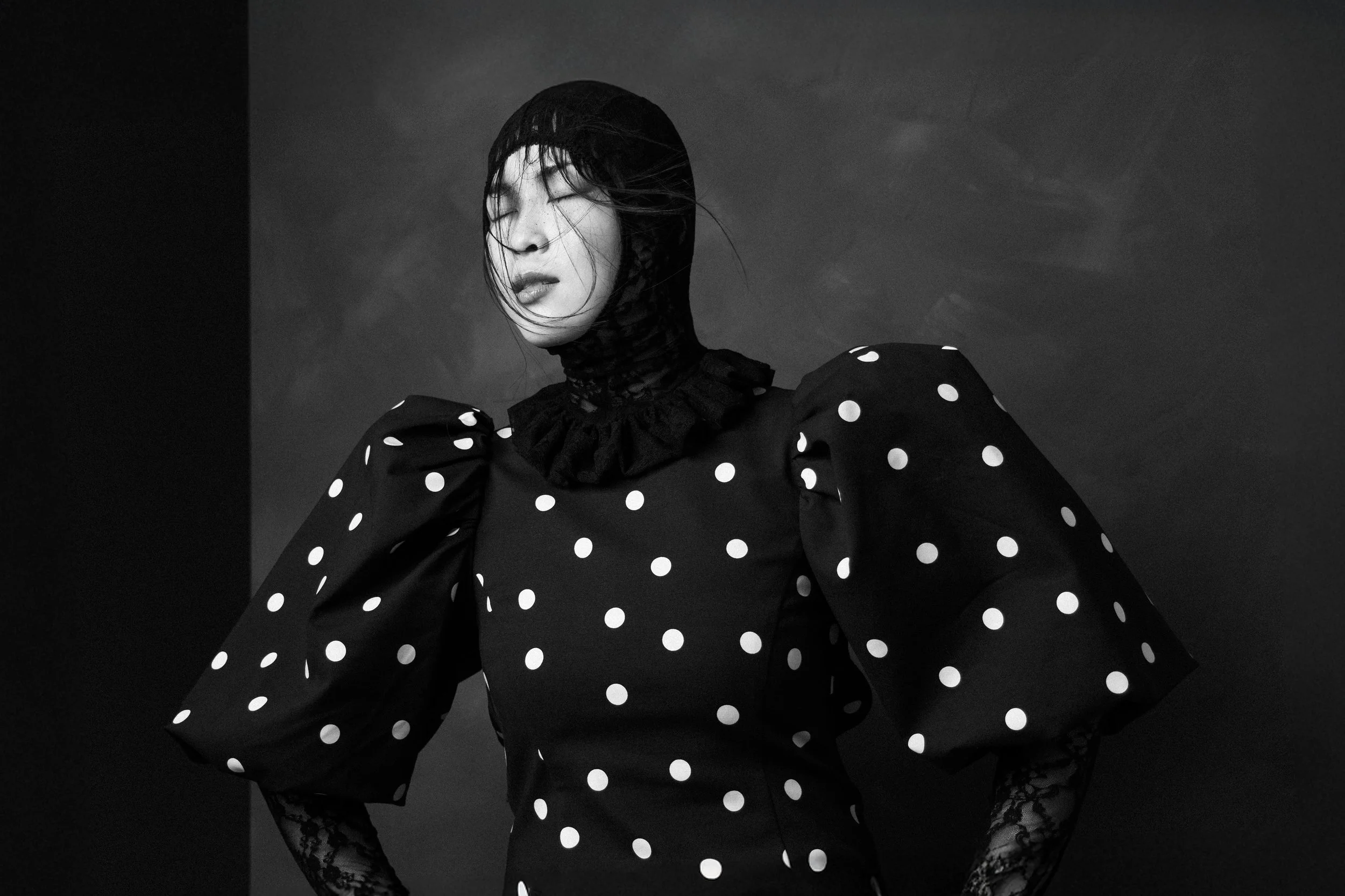 A woman wearing a polka dot dress with puffed sleeves, lace gloves, and a lace collar, with her eyes closed and her hair windblown against a dark background.