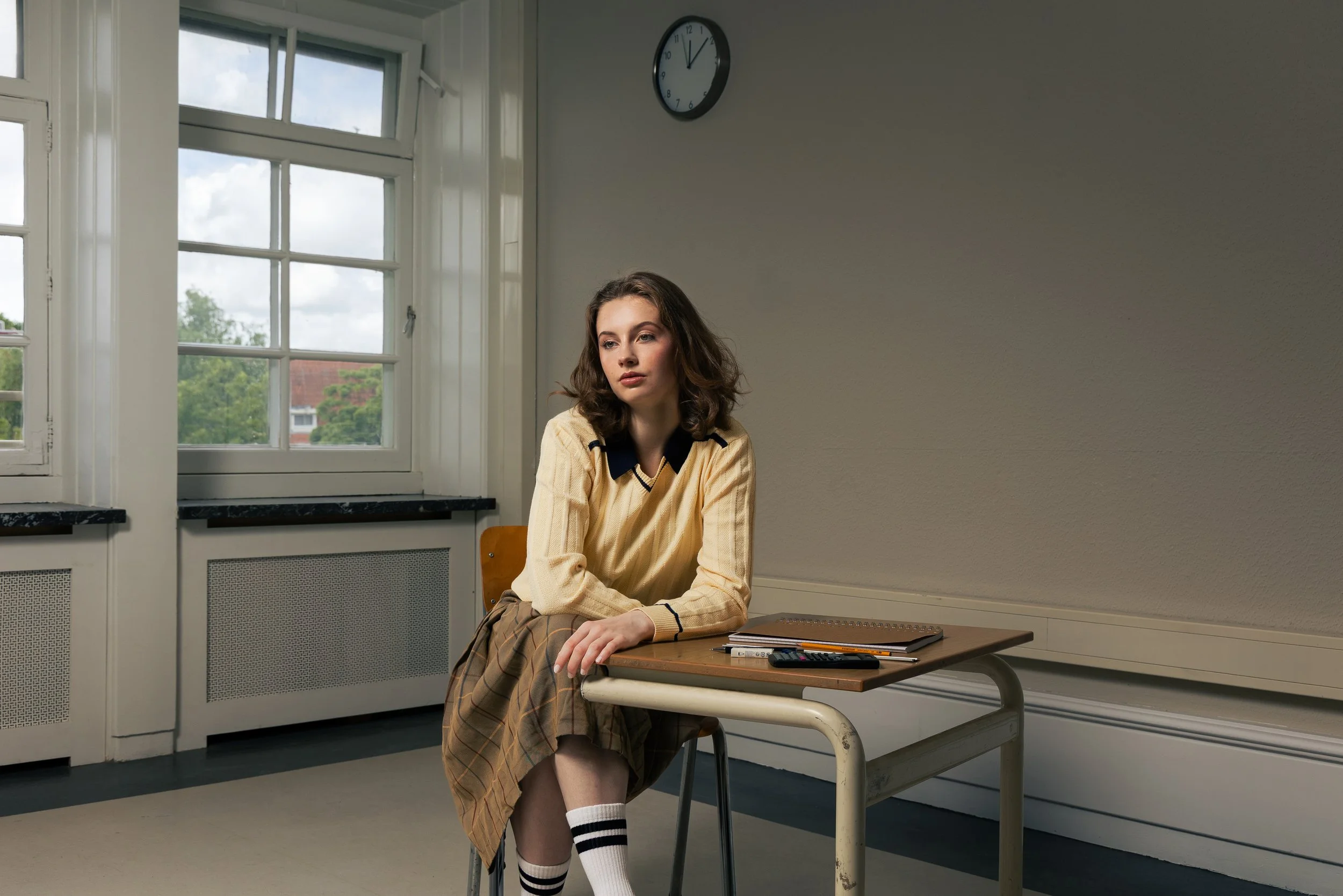 A young woman with brown hair sitting alone at a school desk in a classroom, with a window showing trees outside and a clock on the wall.
