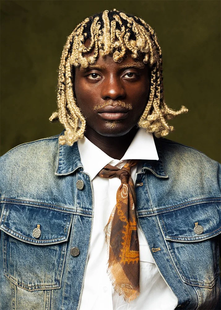 A person with blond twisted hair, dark skin, and a serious expression, wearing a denim jacket over a white shirt and a patterned brown scarf, against a dark green background.