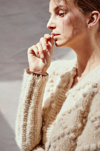 Side profile of a woman with light brown hair wearing a cream-colored, knitted sweater, with her right hand near her chin, looking thoughtfully downward.