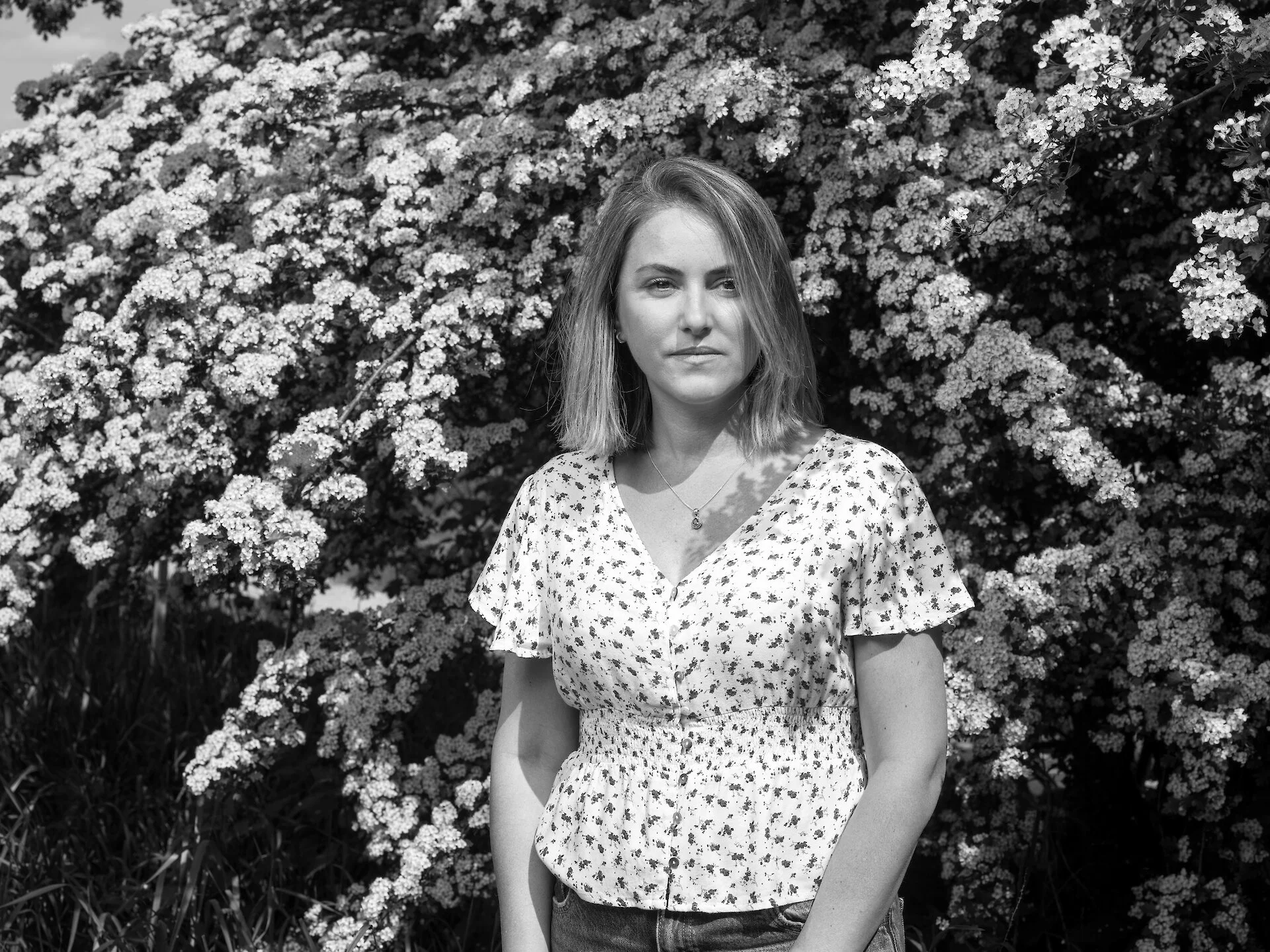 Black and white photo of a young woman with shoulder-length hair standing in front of flowering bushes.