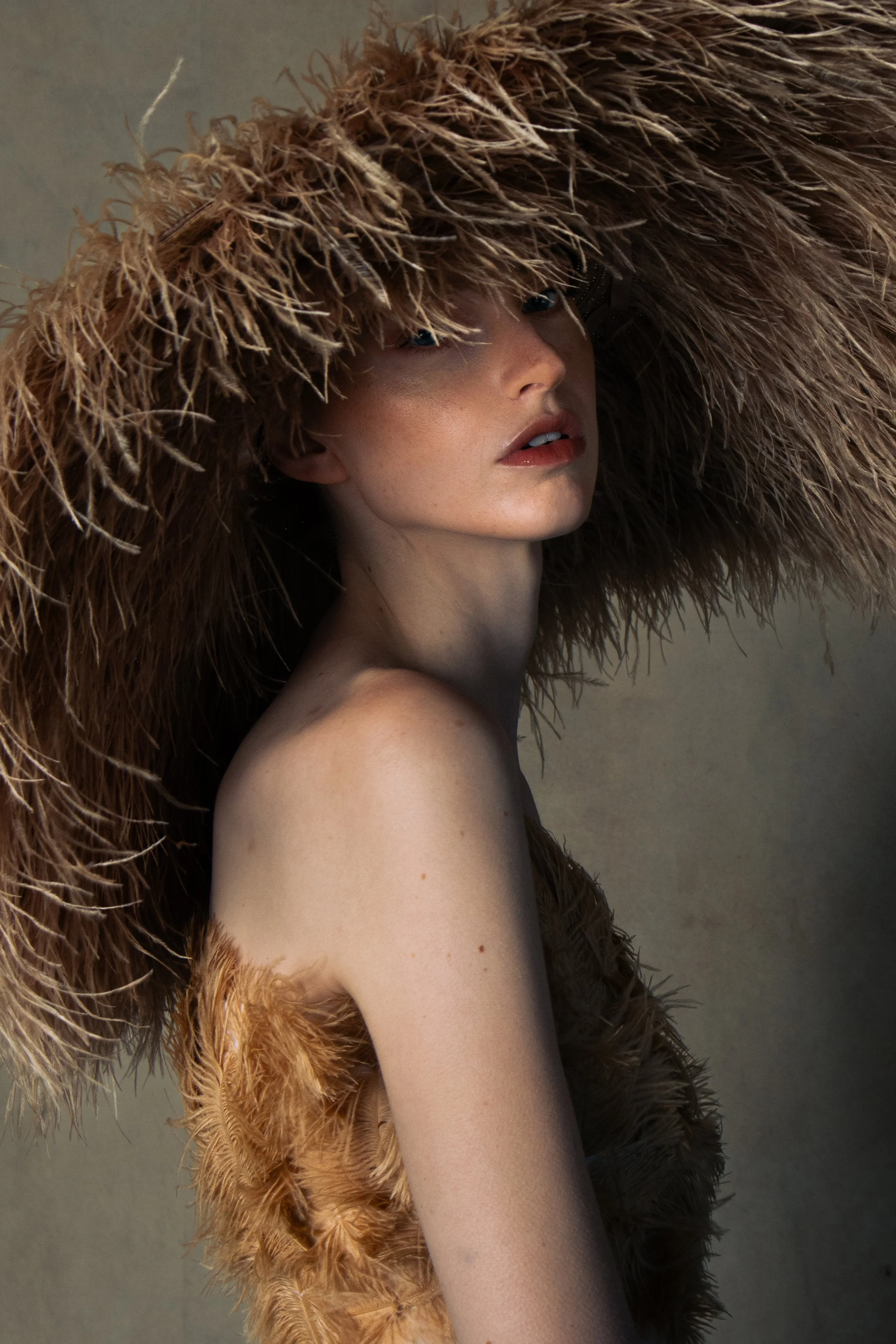 A woman wearing a large, fluffy brown hat and a feathered garment, posing with her shoulder exposed against a neutral background.