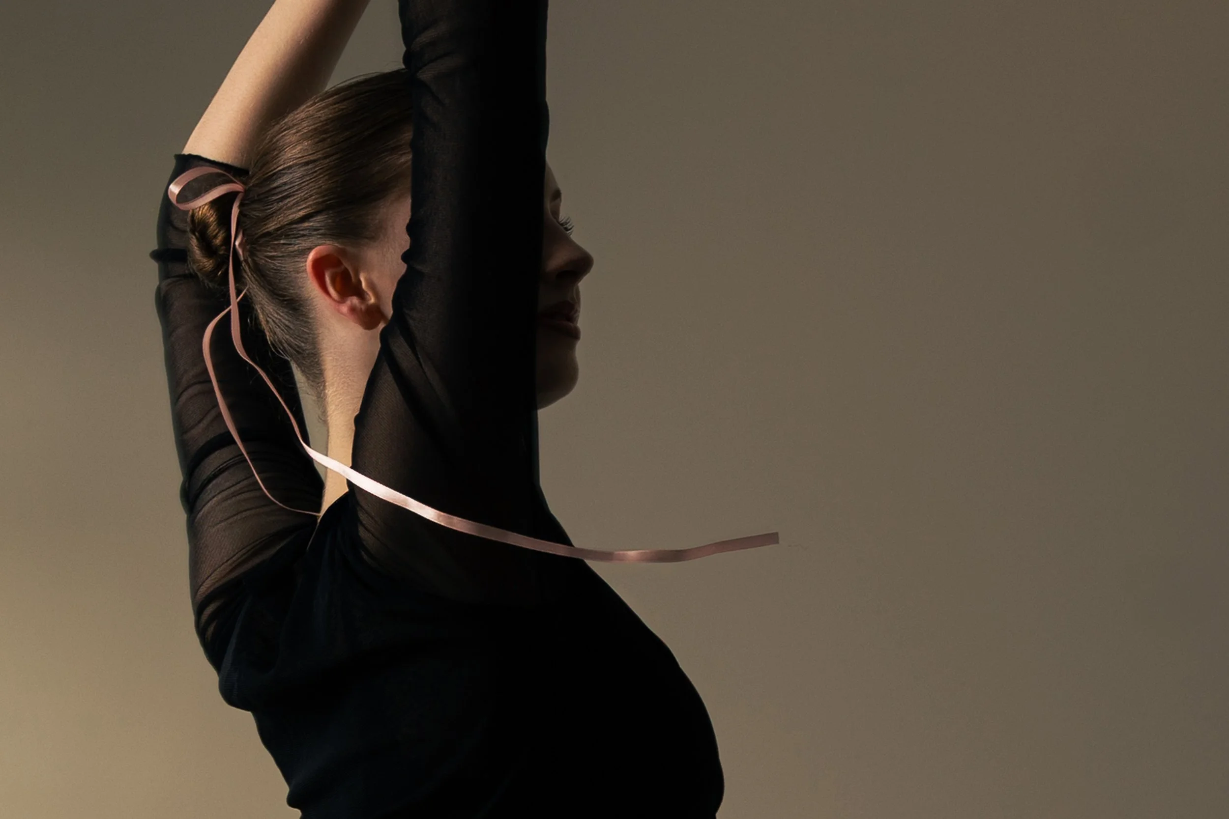 A woman practicing ballet or dance with her arms raised above her head, wearing a black sheer long-sleeved top and a light pink ribbon in her hair, against a plain background.