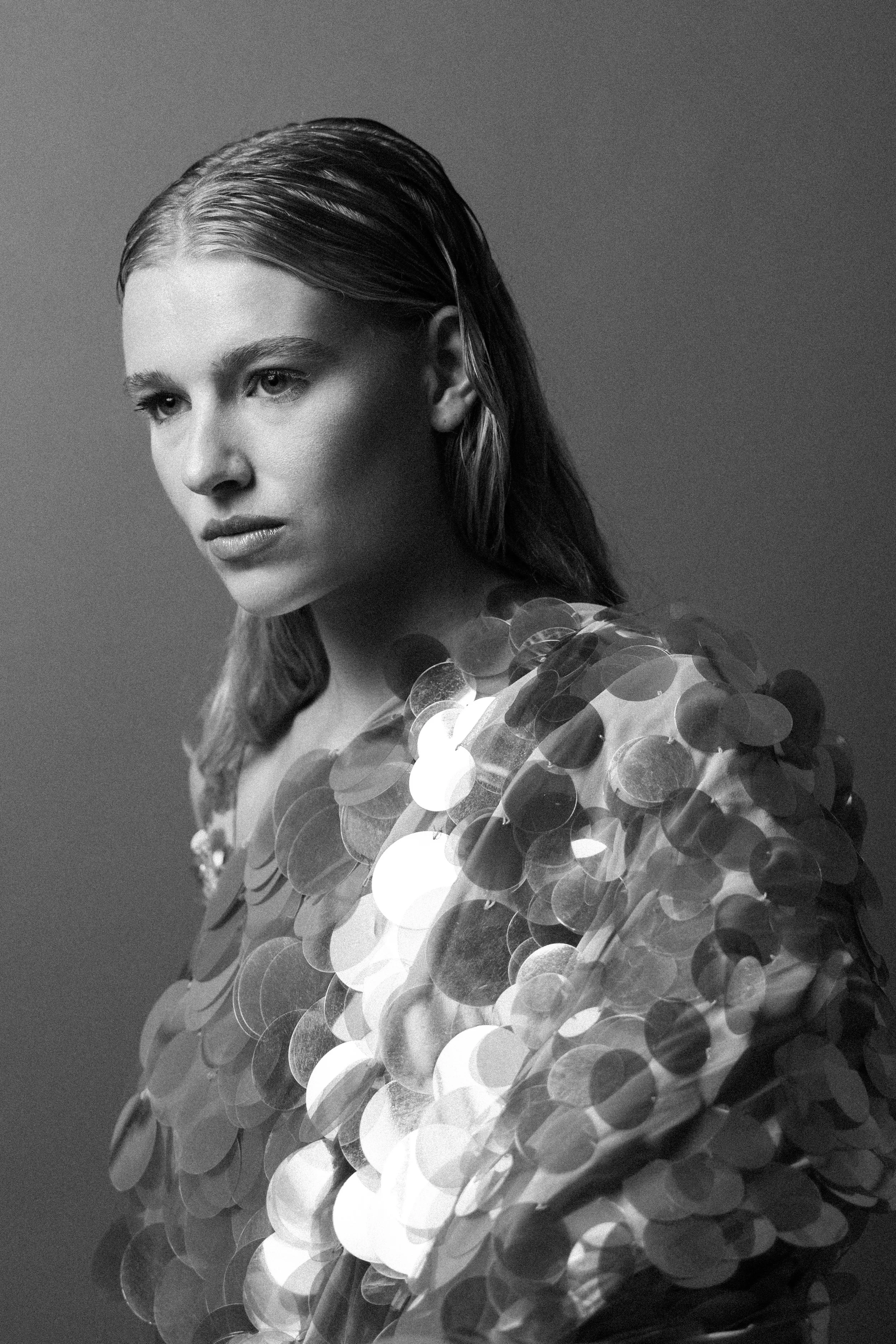 A black-and-white portrait of a woman with long, wet-looking hair, wearing a shiny, reflective sequin dress, against a plain background.