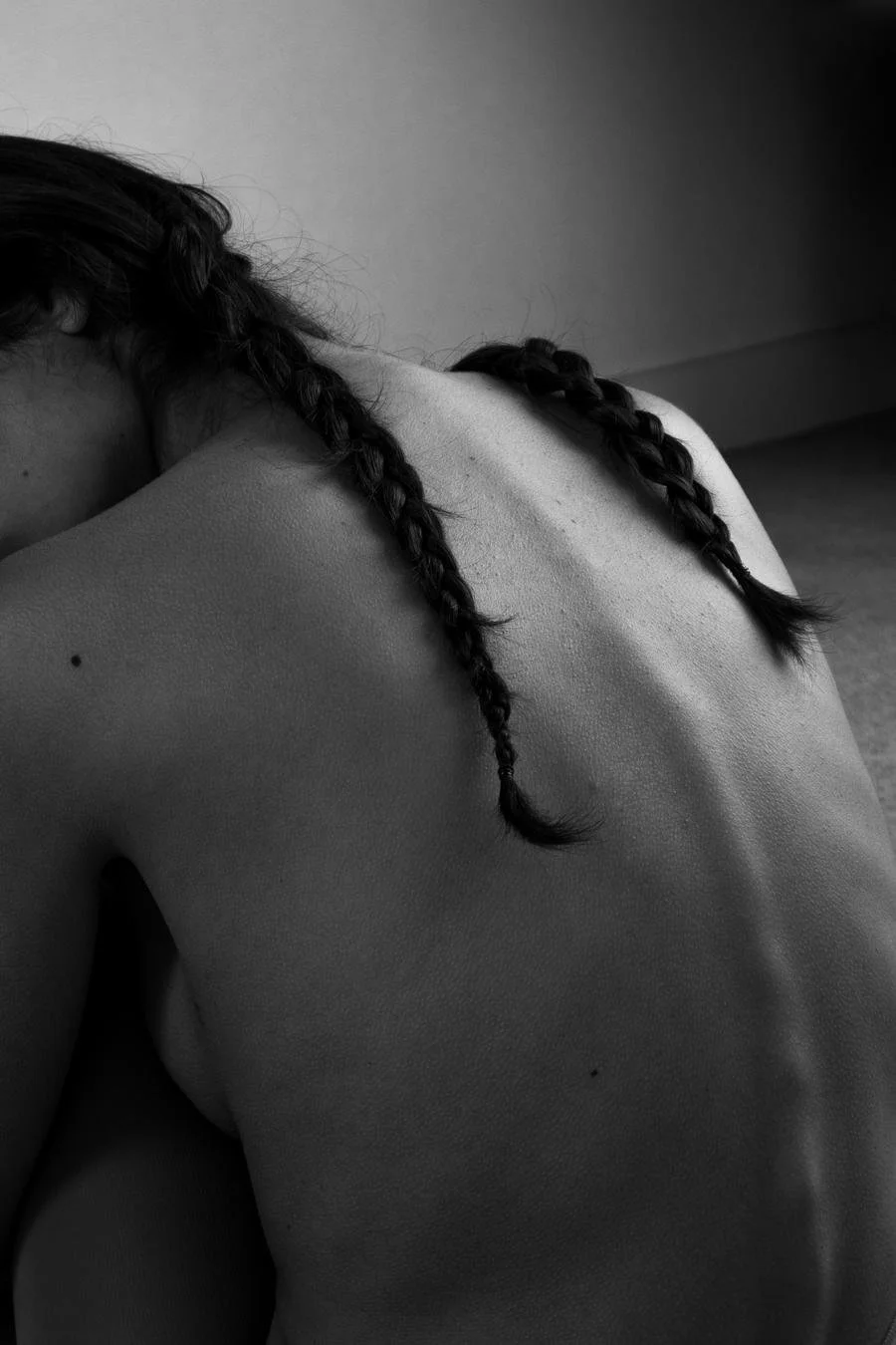 Close-up of a woman's bare back with two braided pigtails, black and white photo, soft lighting, minimal background.