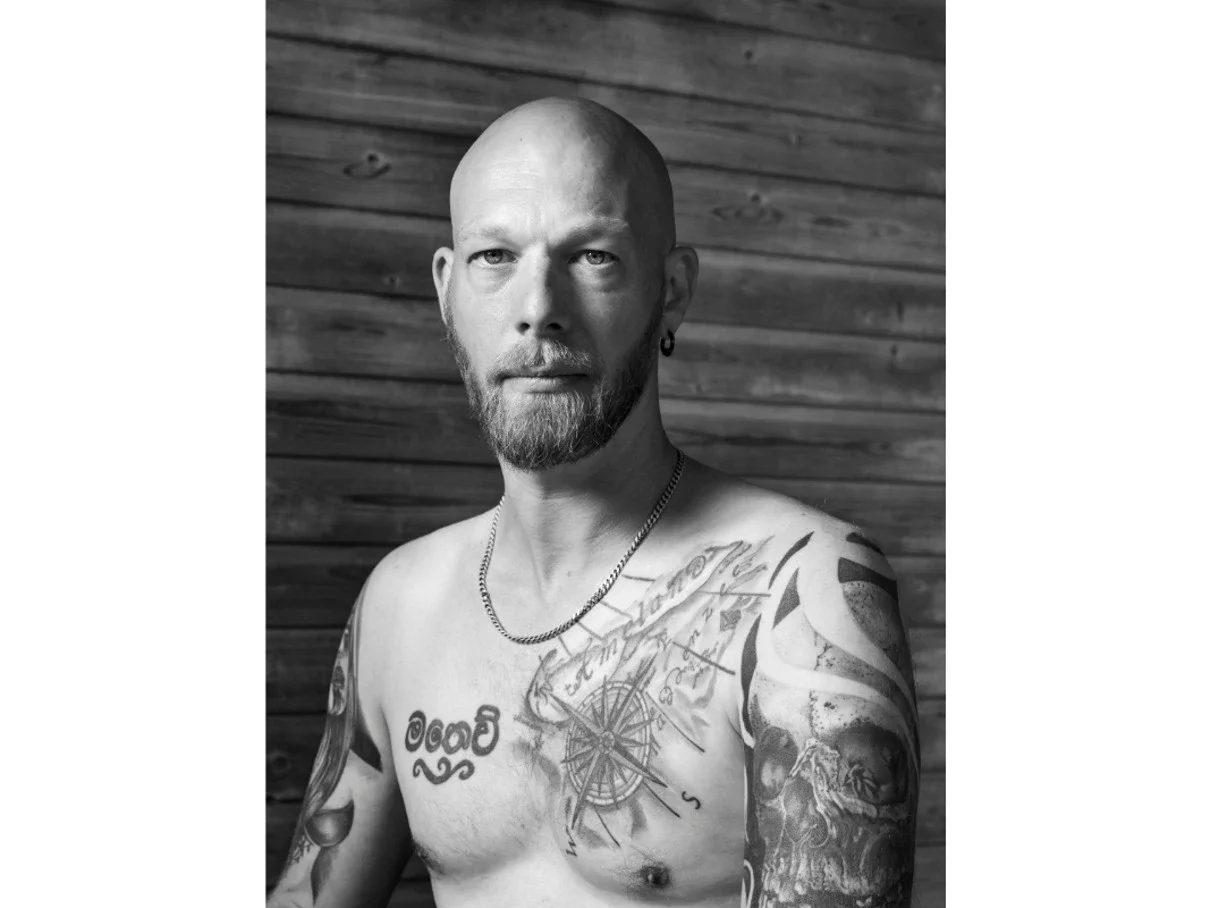 Black and white portrait of a shirtless man with tattoos on his chest and arms, a beard, earring, and chain necklace, set against a wooden background.