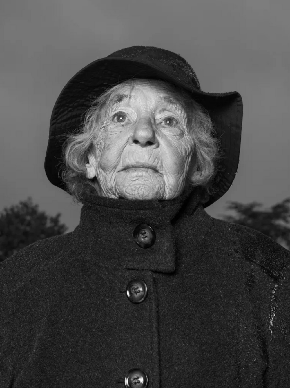 Black and white photo of an elderly woman wearing a wide-brimmed hat and a dark coat with large buttons, looking upward with a serious expression, outdoors with trees in the background.